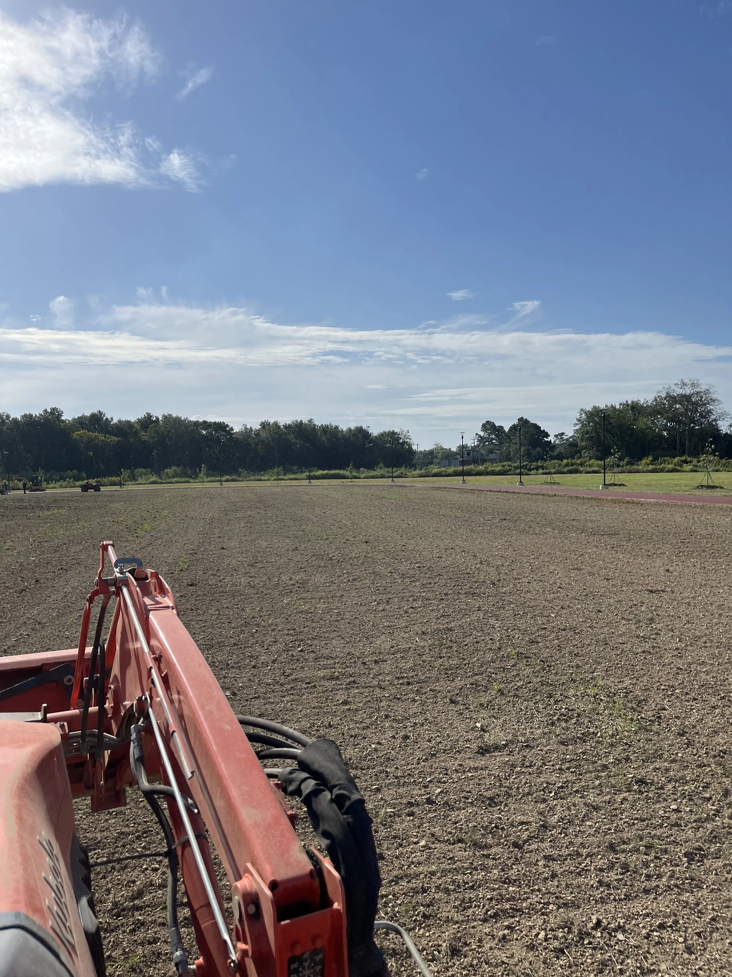 View of a farm field with soil prepared for planting, a tractor or farming equipment in the foreground, a paved pathway, and a line of trees in the distance under a mostly clear blue sky.