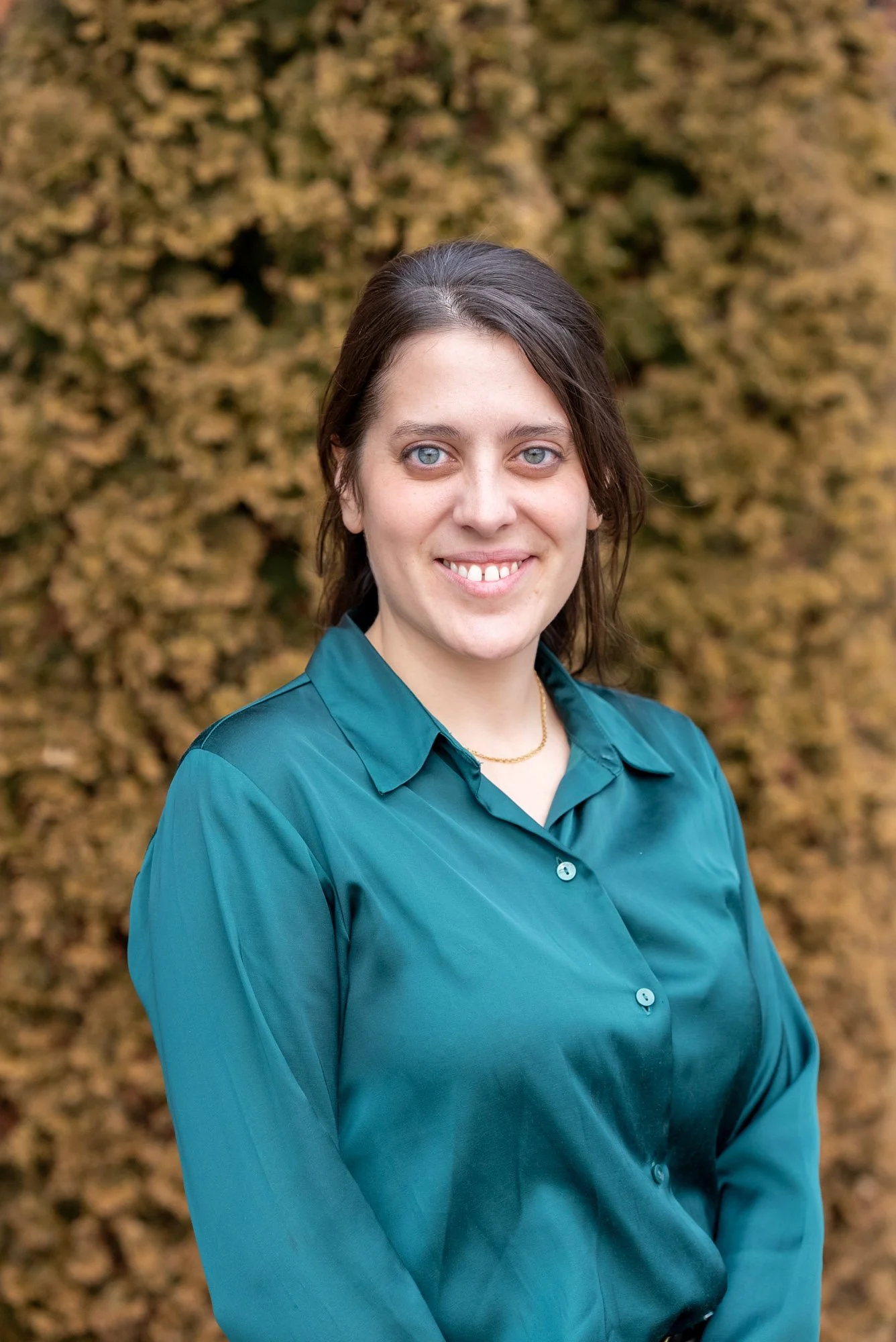 A woman with blue eyes and dark brown hair in a teal blouse, standing outdoors in front of autumn-colored foliage.