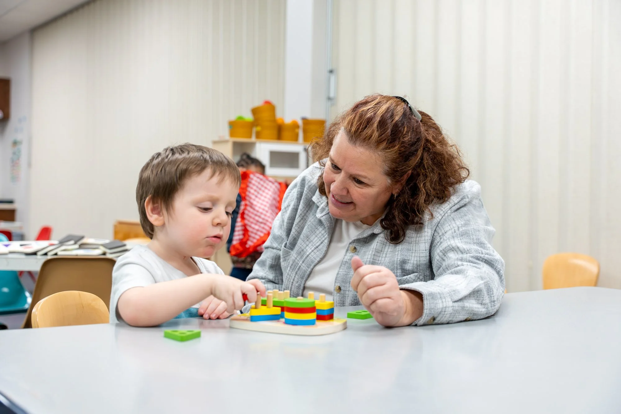 A woman and a young boy playing a board game with colorful pieces at a table in a classroom or activity room.