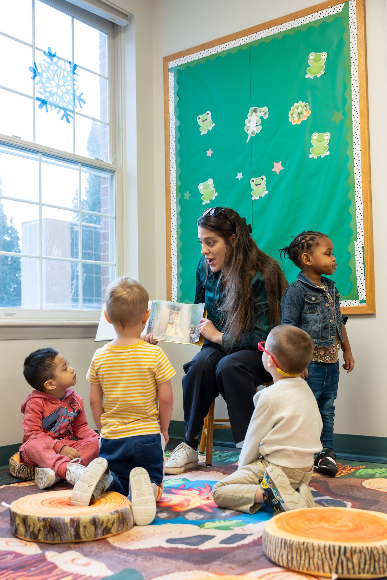 A classroom scene with a woman reading a book to young children. The children are sitting on a colorful rug on tree trunk-shaped cushions. There is a bulletin board with frog and plant decorations, and a window with a snowflake hanging decoration.