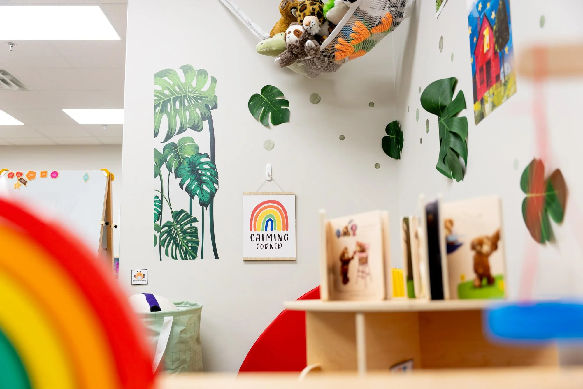A corner of a children's room with tropical leaf wall decals, a sign that says 'Calming Corner', stuffed animal toys in a mesh storage bin, and shelves with books and toys, viewed through an out-of-focus rainbow-colored element.