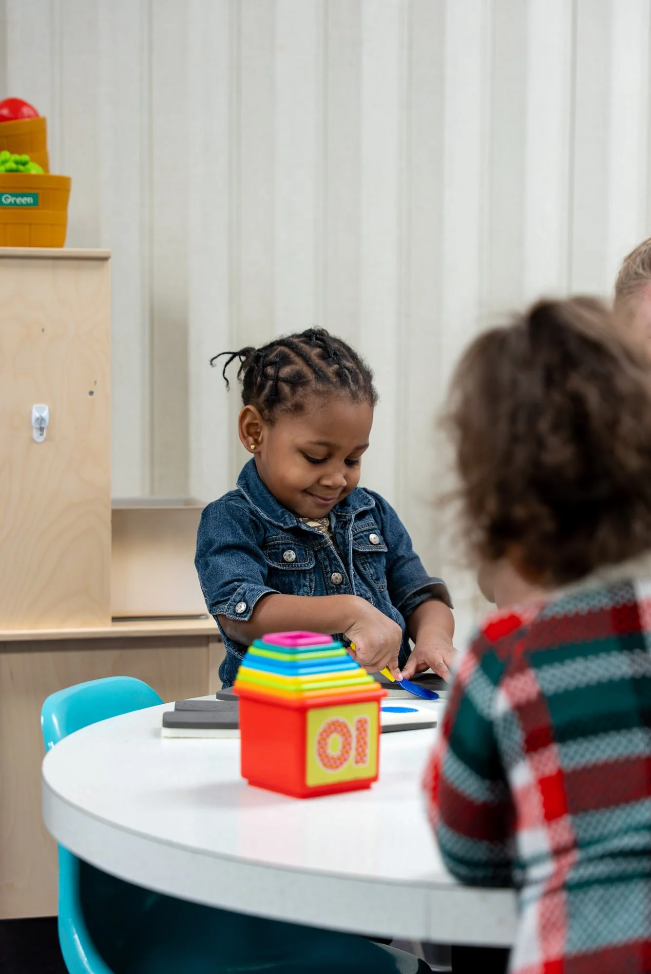 A young girl with braided hair and earrings is playing with colorful stacking toys at a table in a classroom, with other children partially visible.