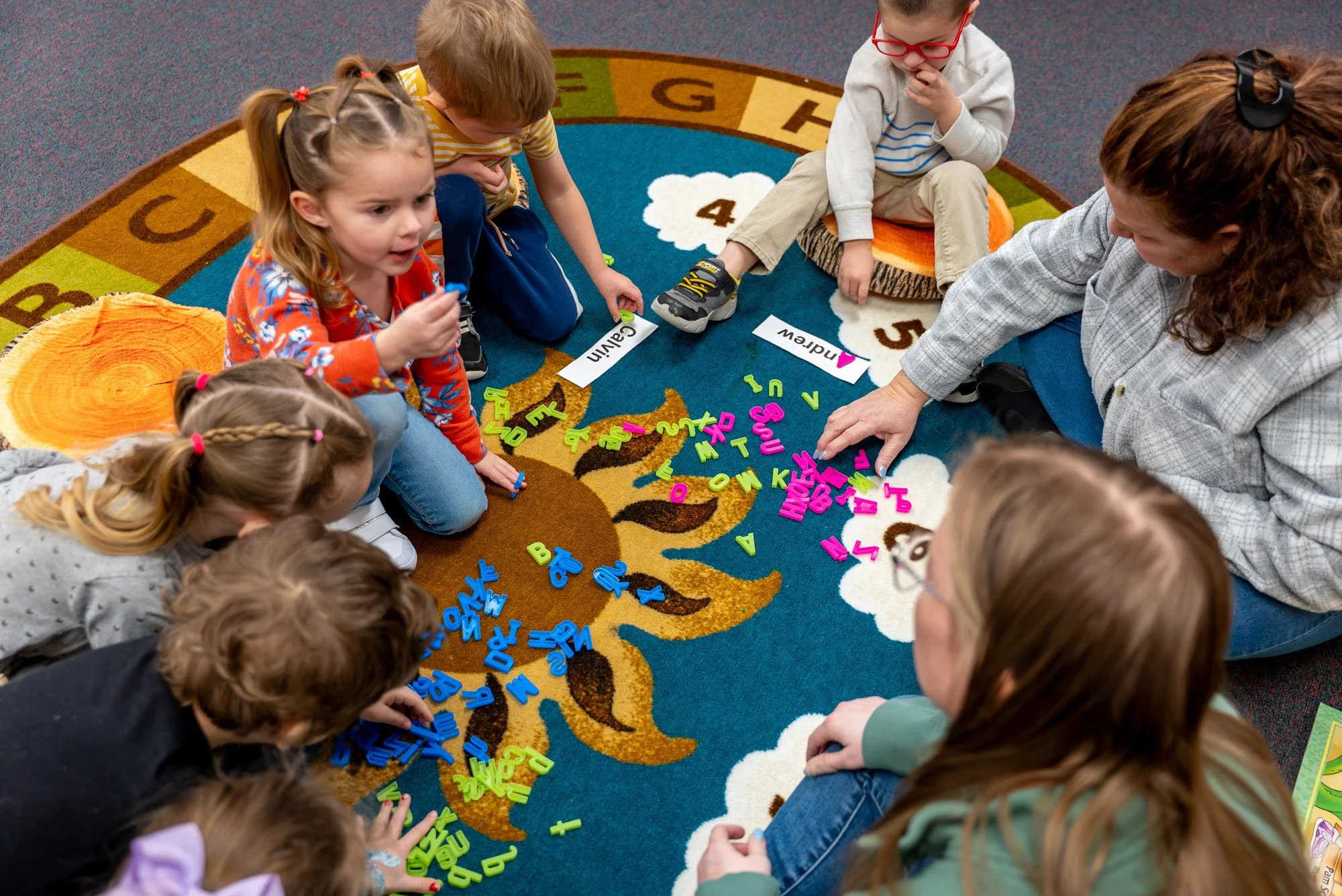 A teacher and a group of children sitting on a colorful rug with letters and numbers, engaging in a learning activity with alphabet foam letters and flashcards. The scene appears to be in a classroom.