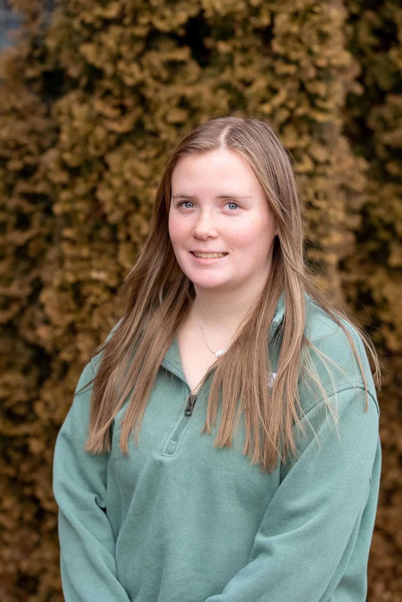 A young woman with long, light brown hair and blue eyes smiling at the camera, standing outside in front of a background of blurred brownish-yellow autumn leaves.