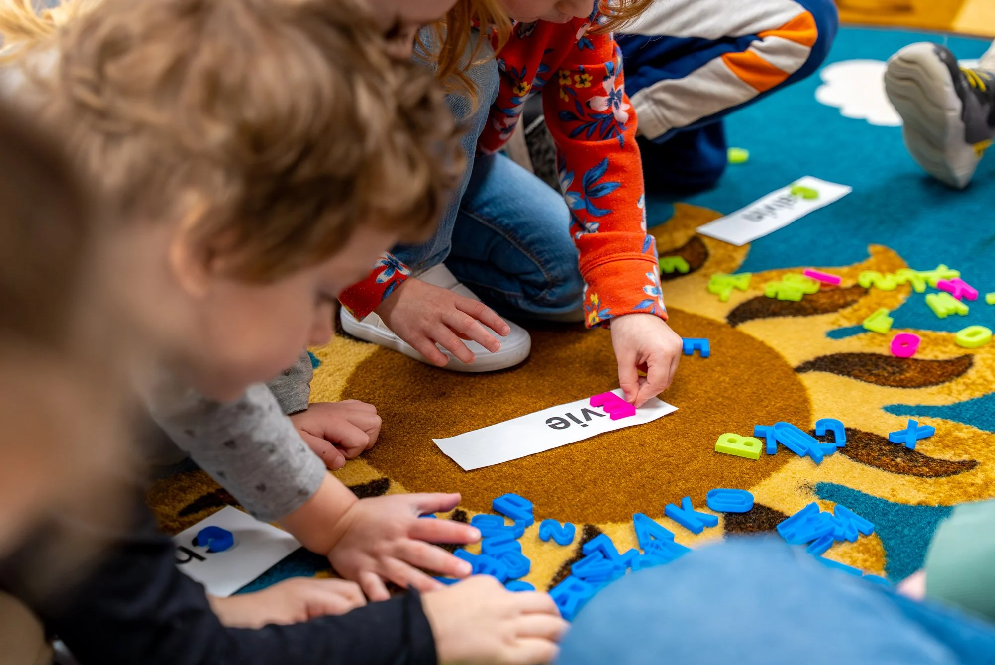 Children sitting on a colorful carpet, playing with magnetic alphabet letters, spelling the word 'VIE' with some colorful letters scattered around.