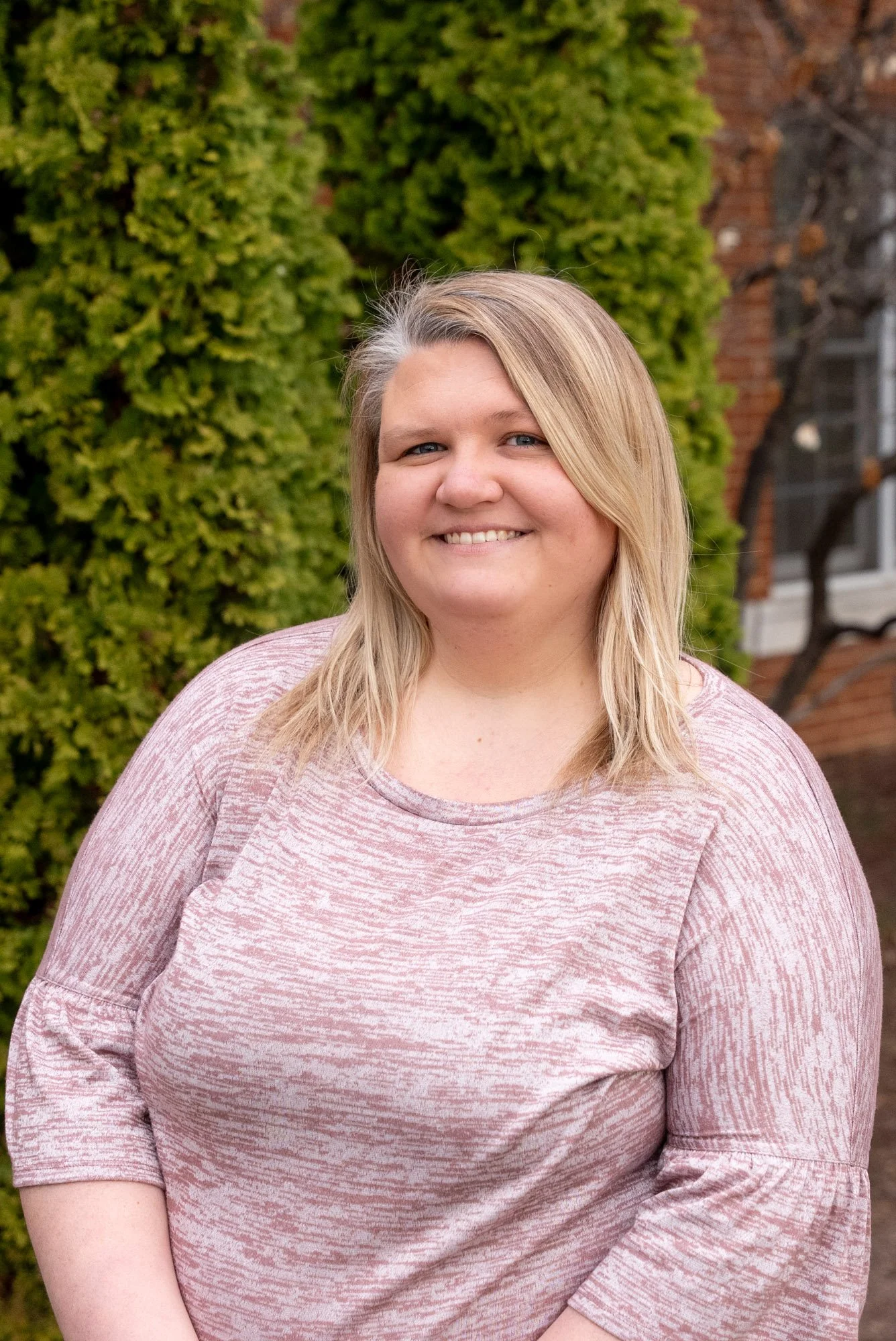 A woman with blonde hair and blue eyes smiling wearing a light pink patterned shirt outdoors, with green bushes and a brick building in the background.