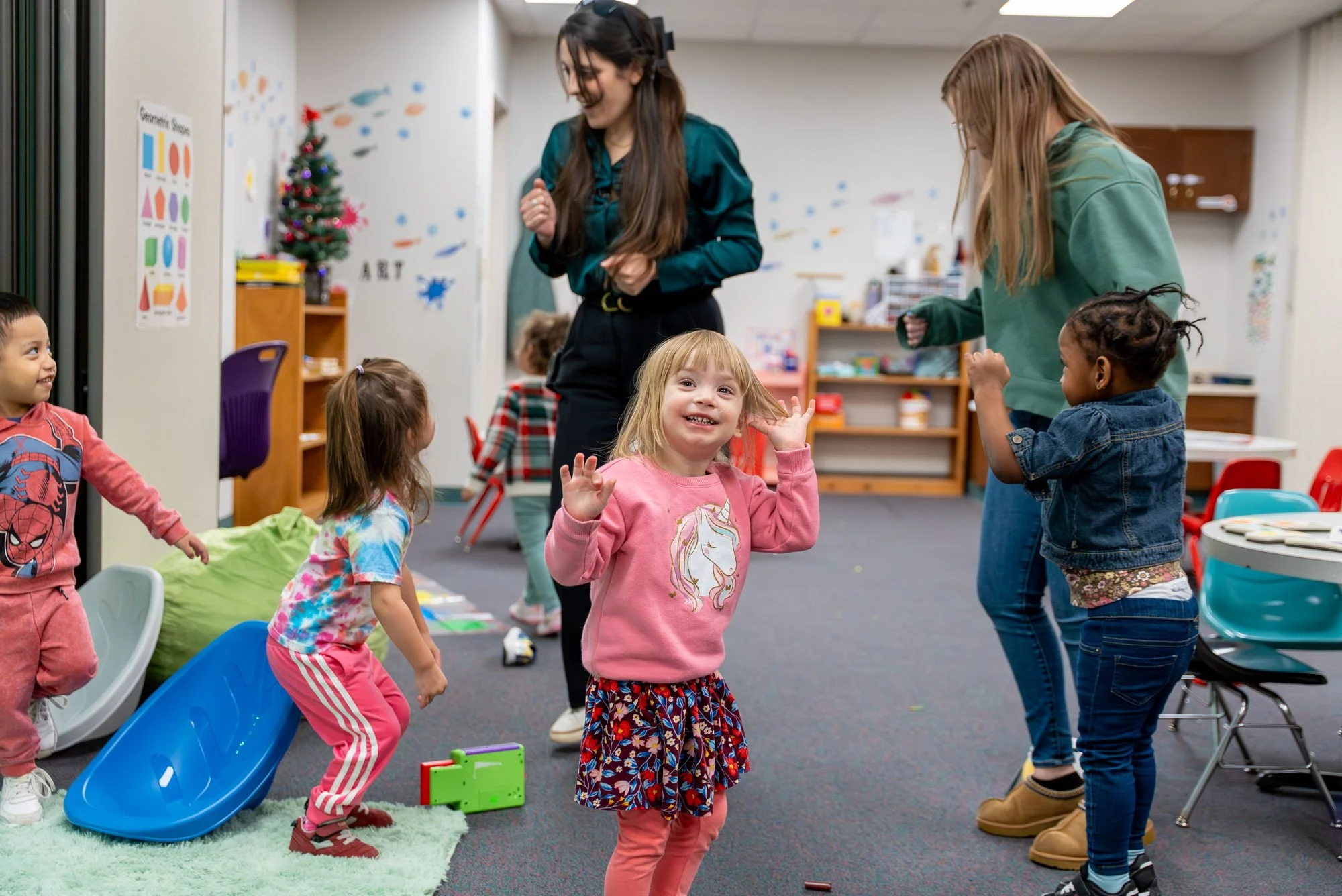 Children playing and laughing in a classroom decorated for Christmas, with two women encouraging the kids, some seated and some standing, engaging in a playful activity.
