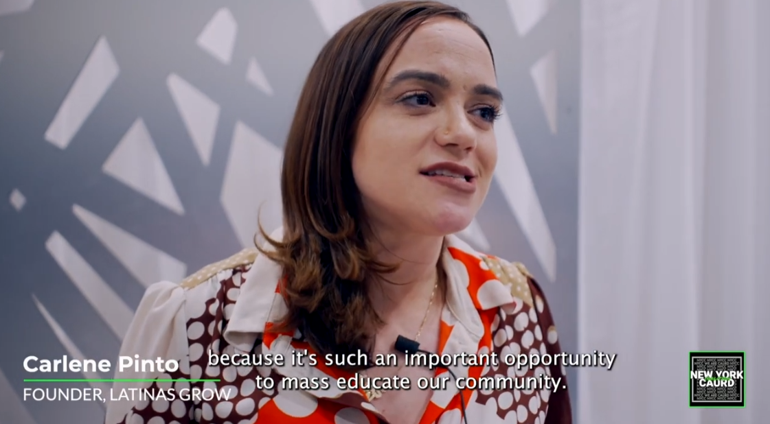 A woman with shoulder-length brown hair speaking during an interview, standing in front of an abstract geometric gray and white background.