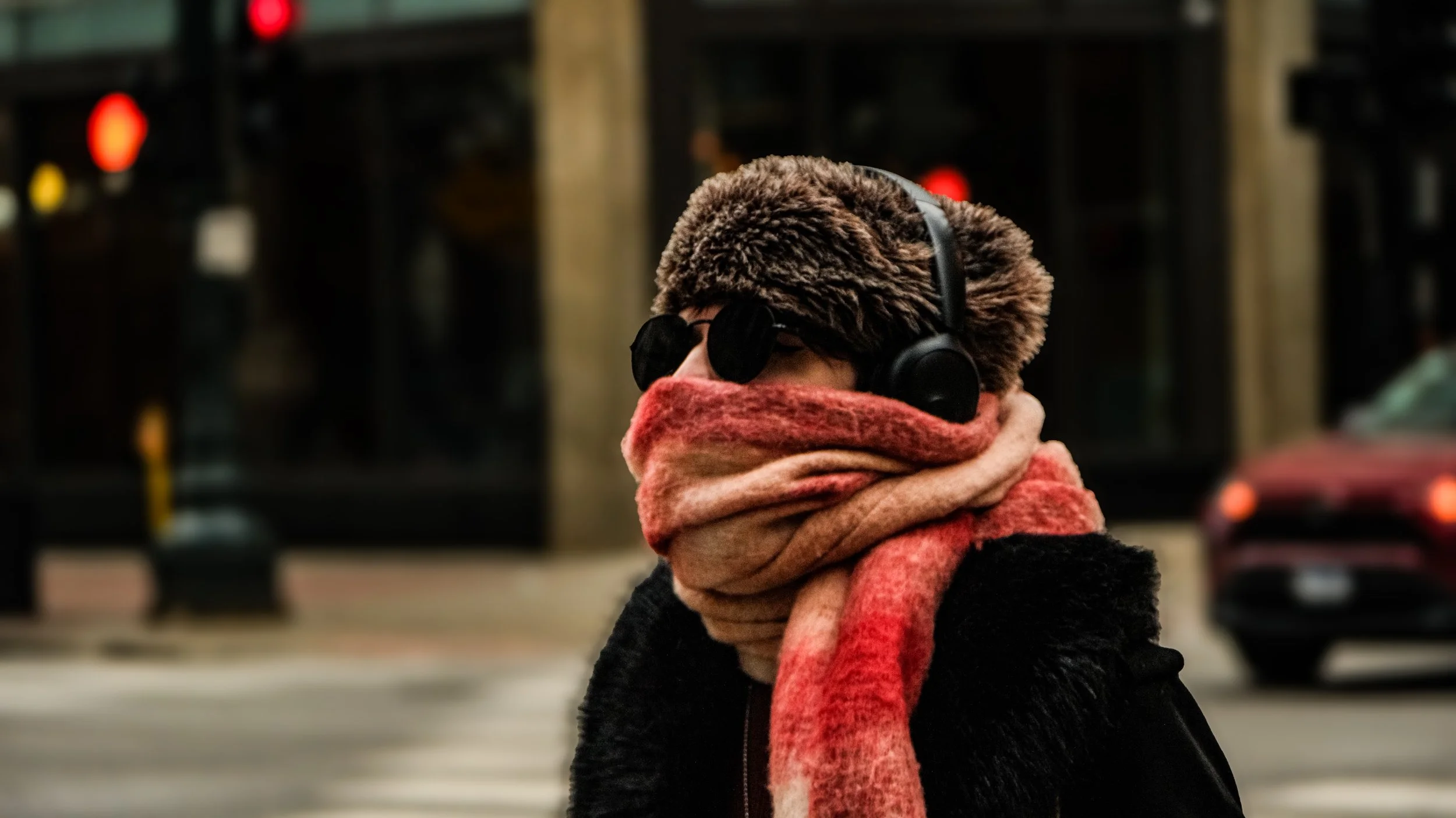 A woman standing on a city street at night, wearing a furry hat, sunglasses, headphones, scarf, and a black coat.