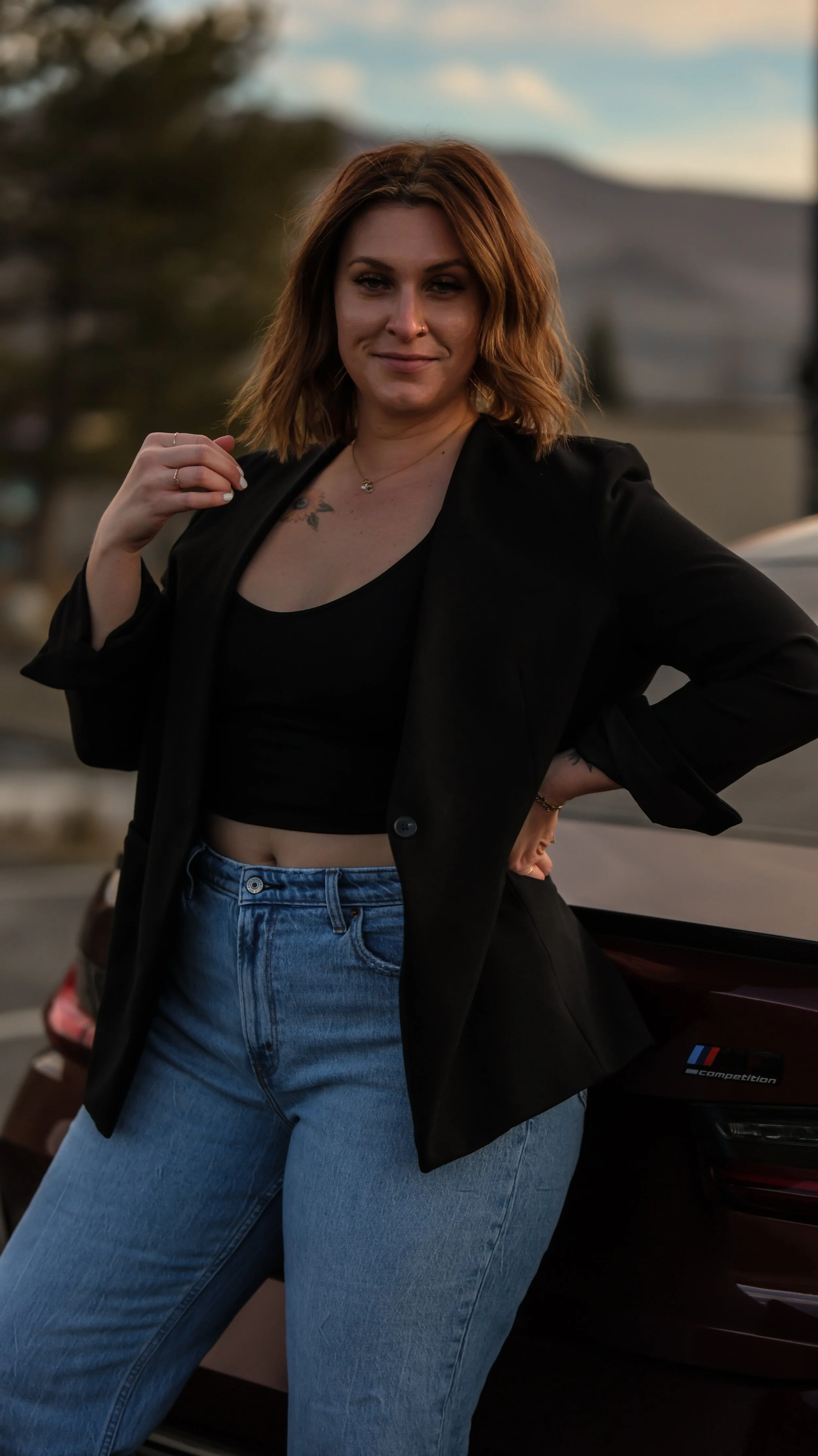 A young woman with shoulder-length auburn hair standing outdoors next to a black car during sunset, wearing a black crop top, black blazer, and high-waisted blue jeans.