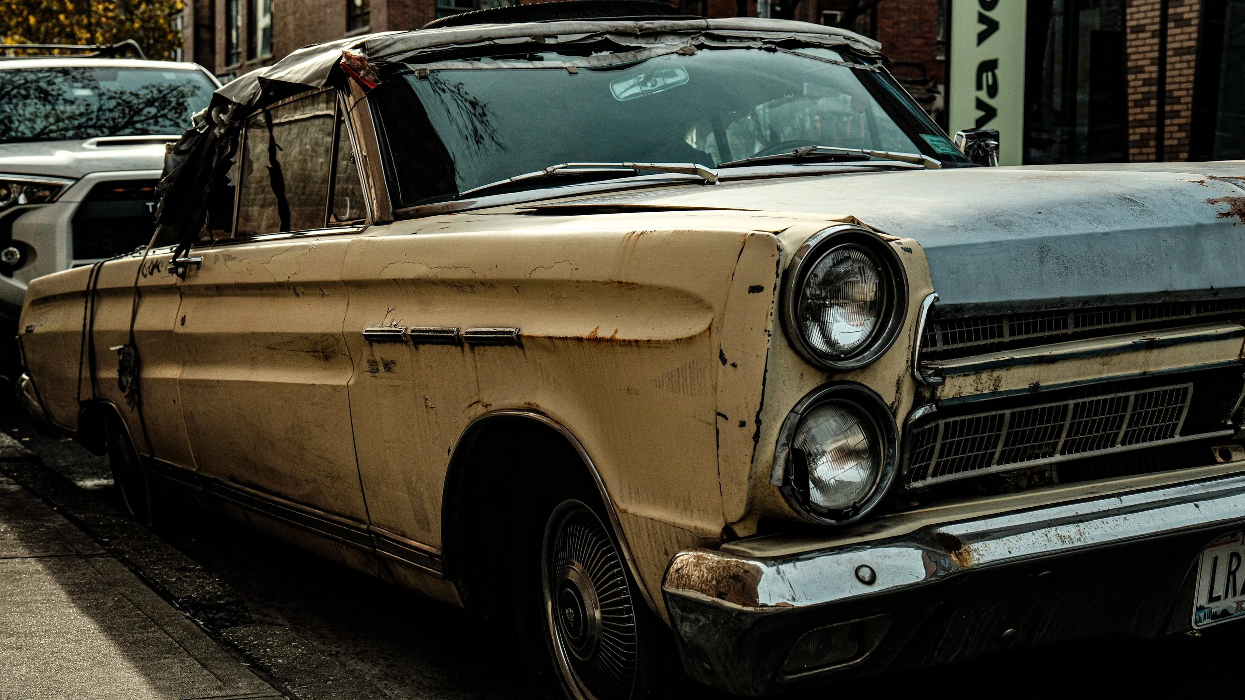 An old, rusty beige car with visible dents, rust spots, and a damaged windshield parked on the side of a street.