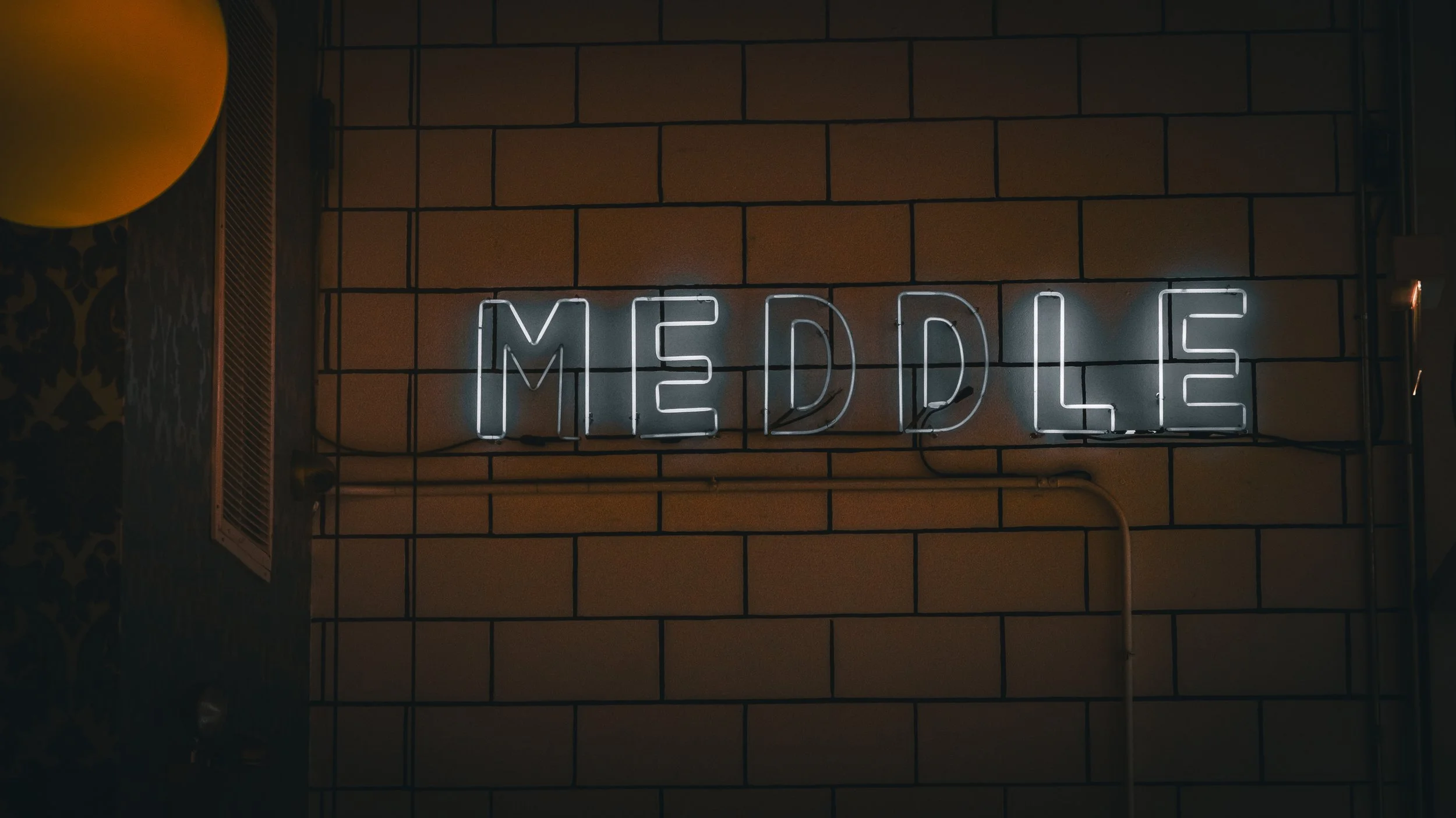 Neon sign reading 'MEDDLE' mounted on a brick wall in a dimly lit interior.