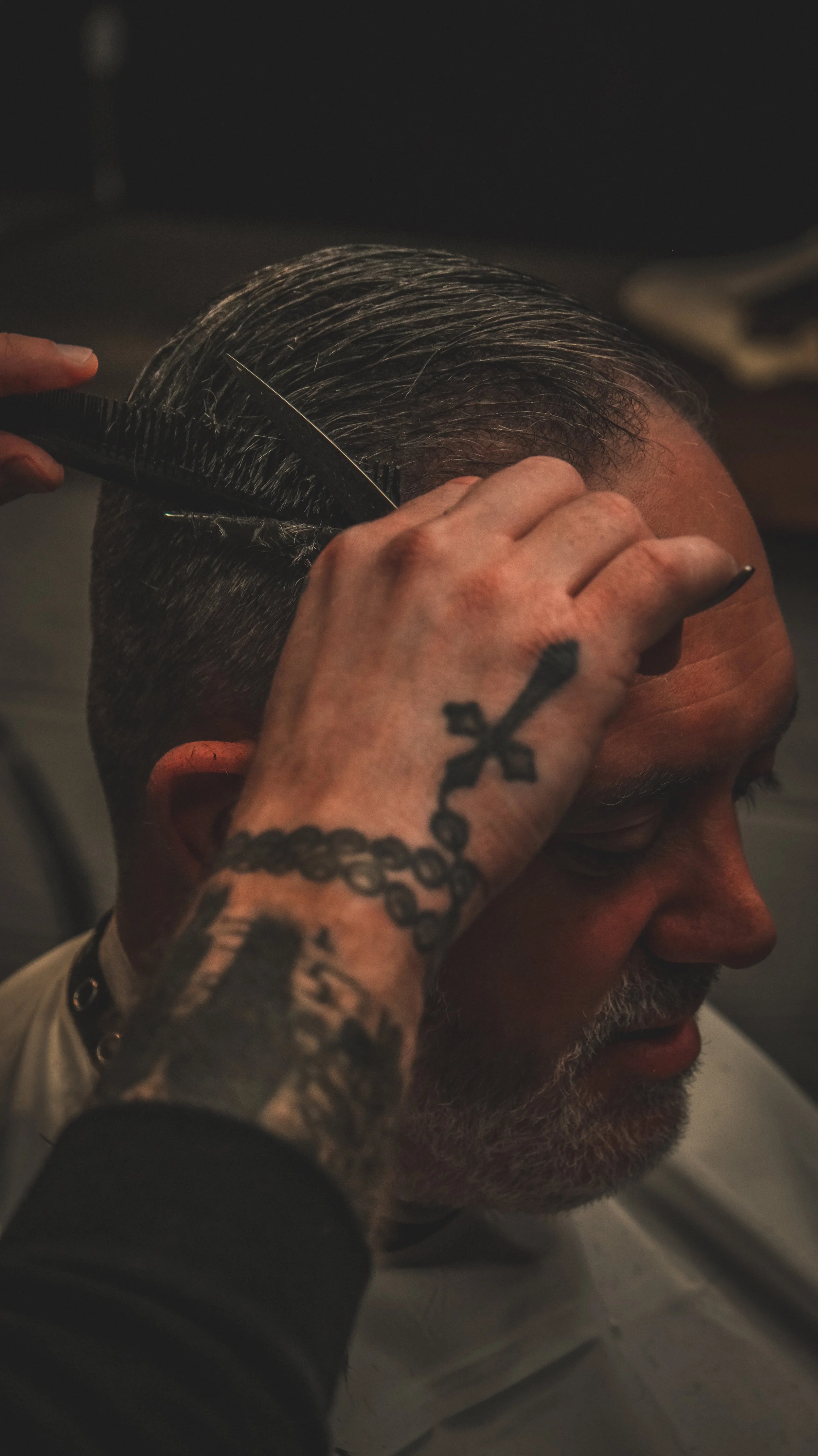 A person getting a haircut, with a barber using scissors and a comb to style the hair, and a tattoo of a cross and chain on their hand.