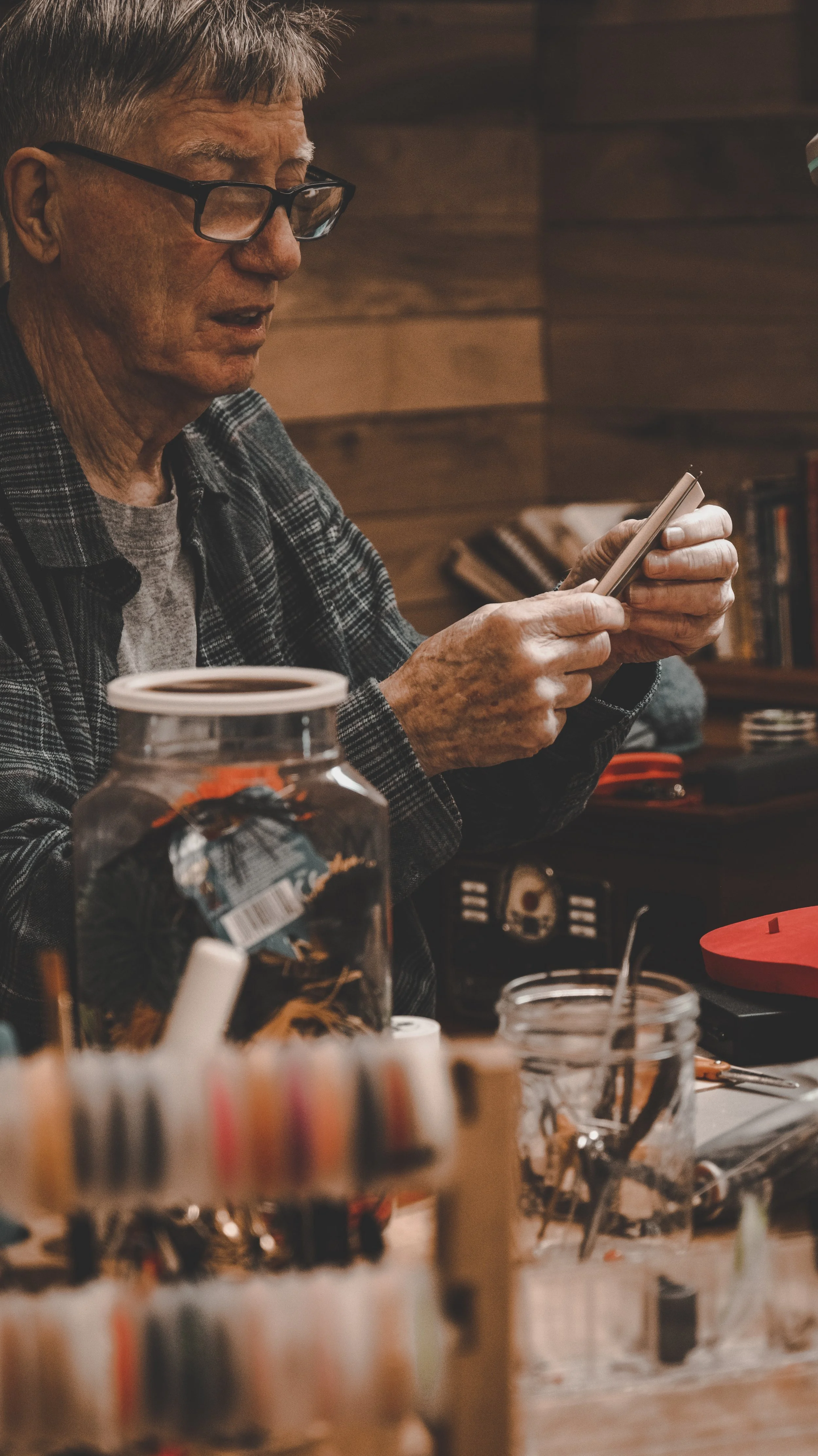 An elderly man with glasses wearing a plaid shirt, looking at his phone in a cozy, wood-paneled room.