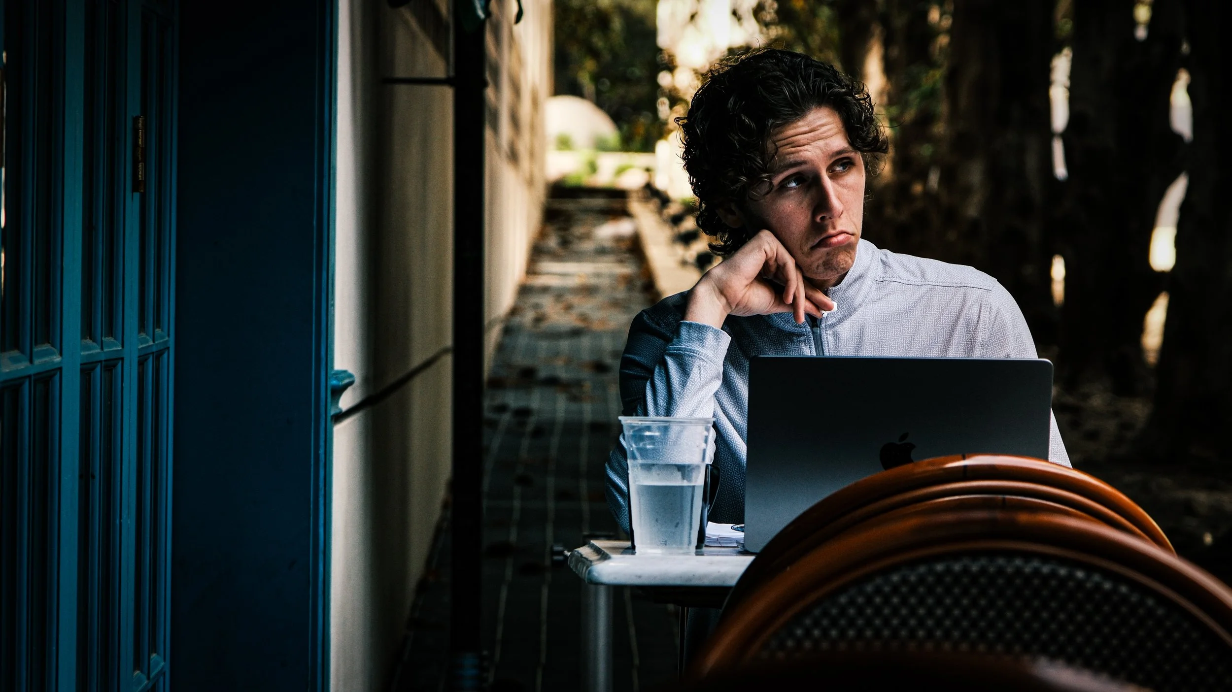 A young man with curly hair sitting outside at a table with a laptop and a glass of water. He appears to be bored or lost in thought, resting his chin on his hand.