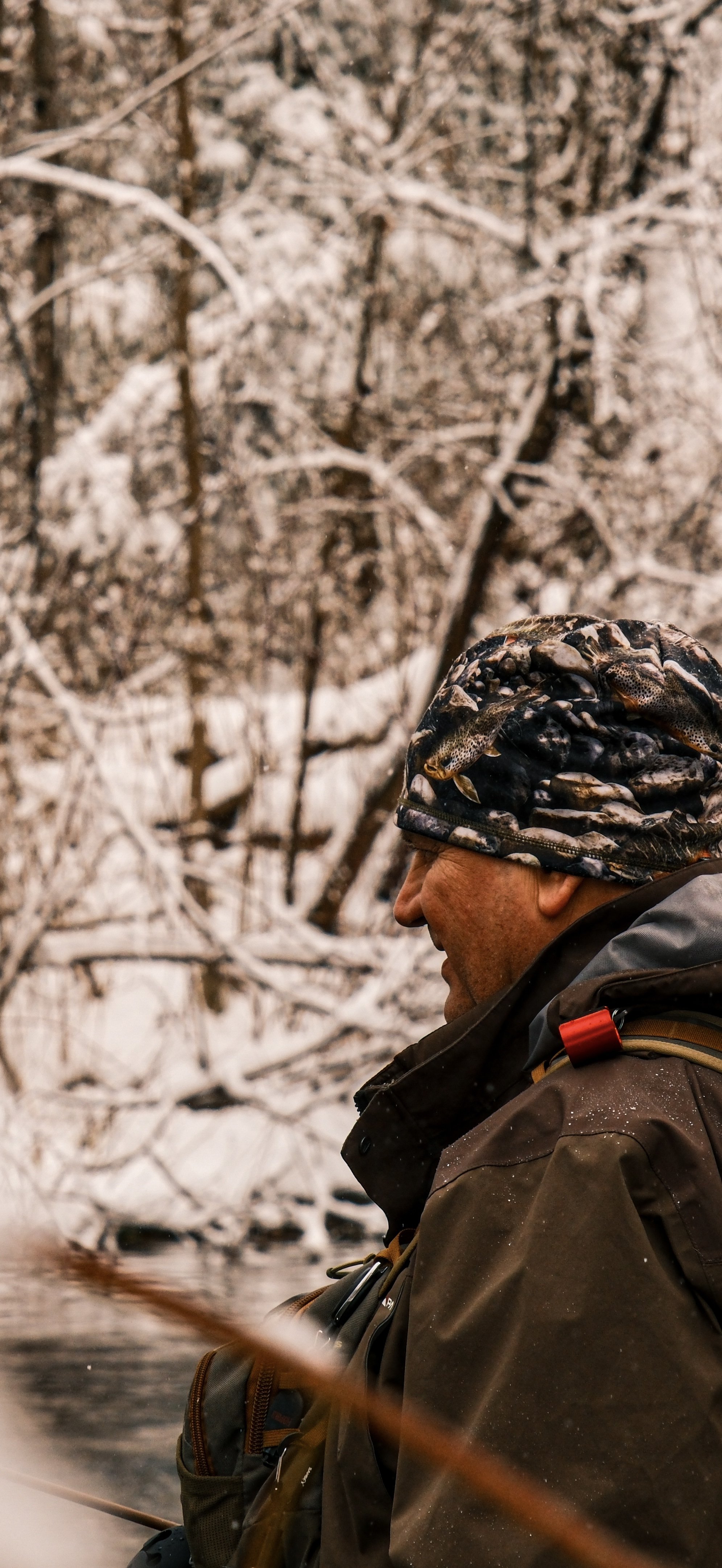A man dressed in outdoor gear, wearing a camouflage hat, sitting outdoors near a snow-covered landscape with trees in the background.