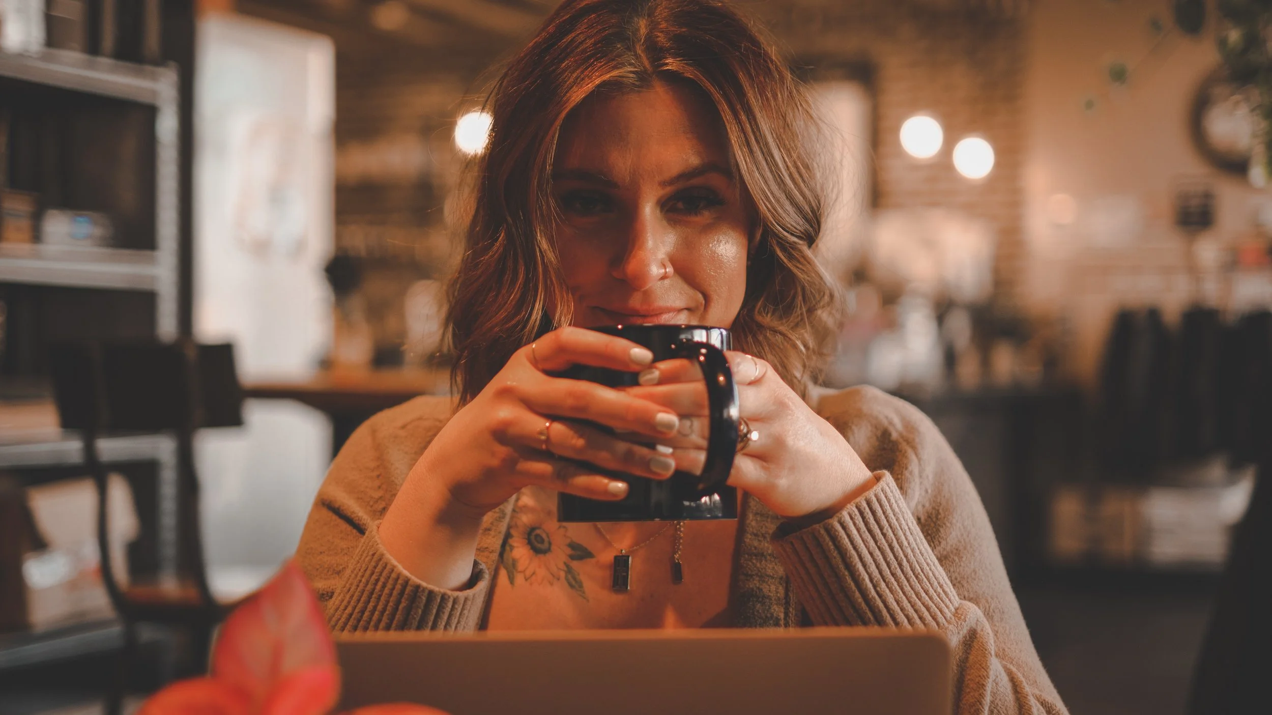 A woman with wavy brown hair, wearing a beige sweater, holding a black coffee mug in a cozy cafe, looking at the camera with a subtle smile.