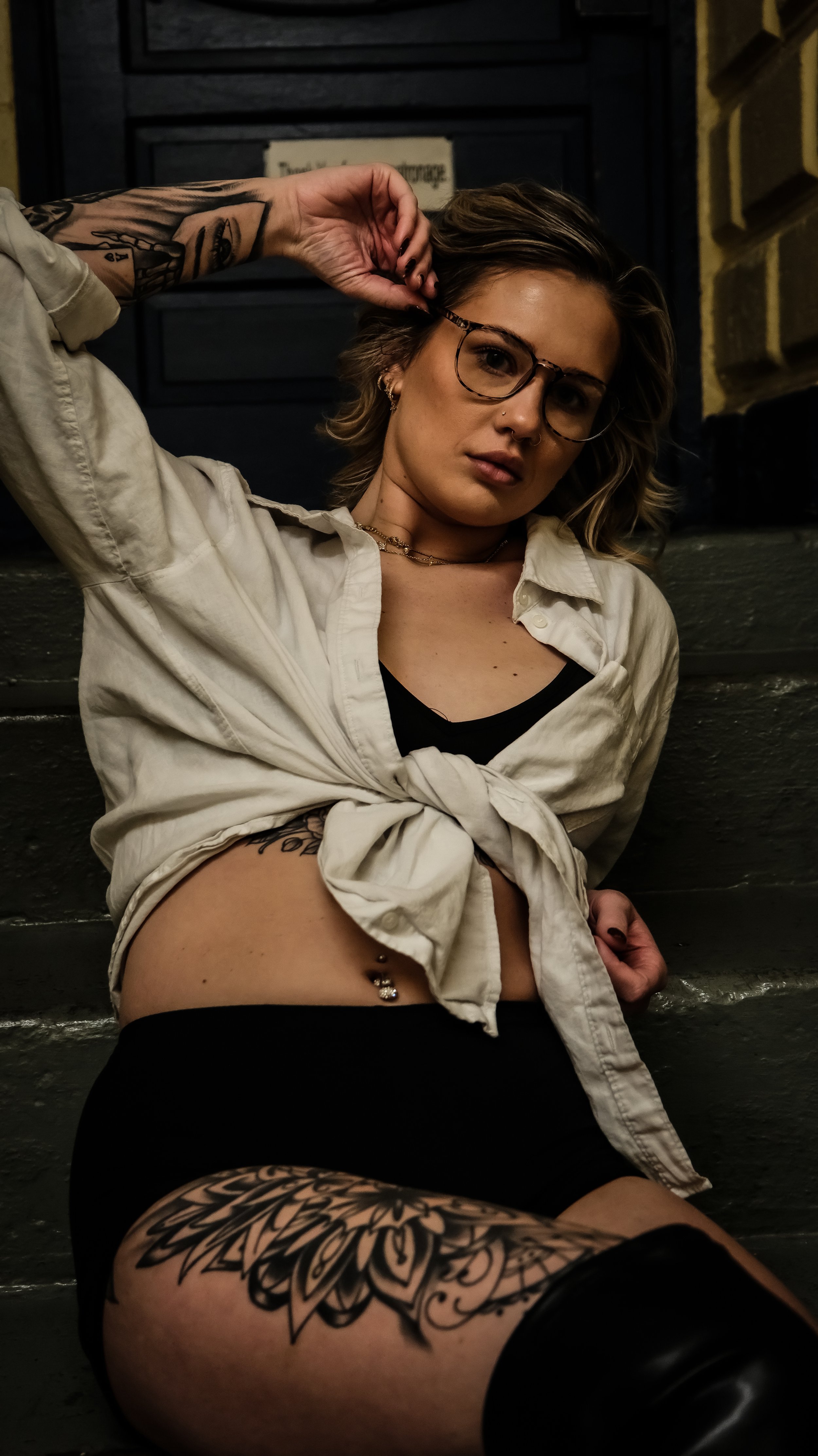 A young woman with tattoos, glasses, and medium-length hair, sitting on stairs against a dark wall and brick, wearing a knotted light-colored shirt and black shorts.