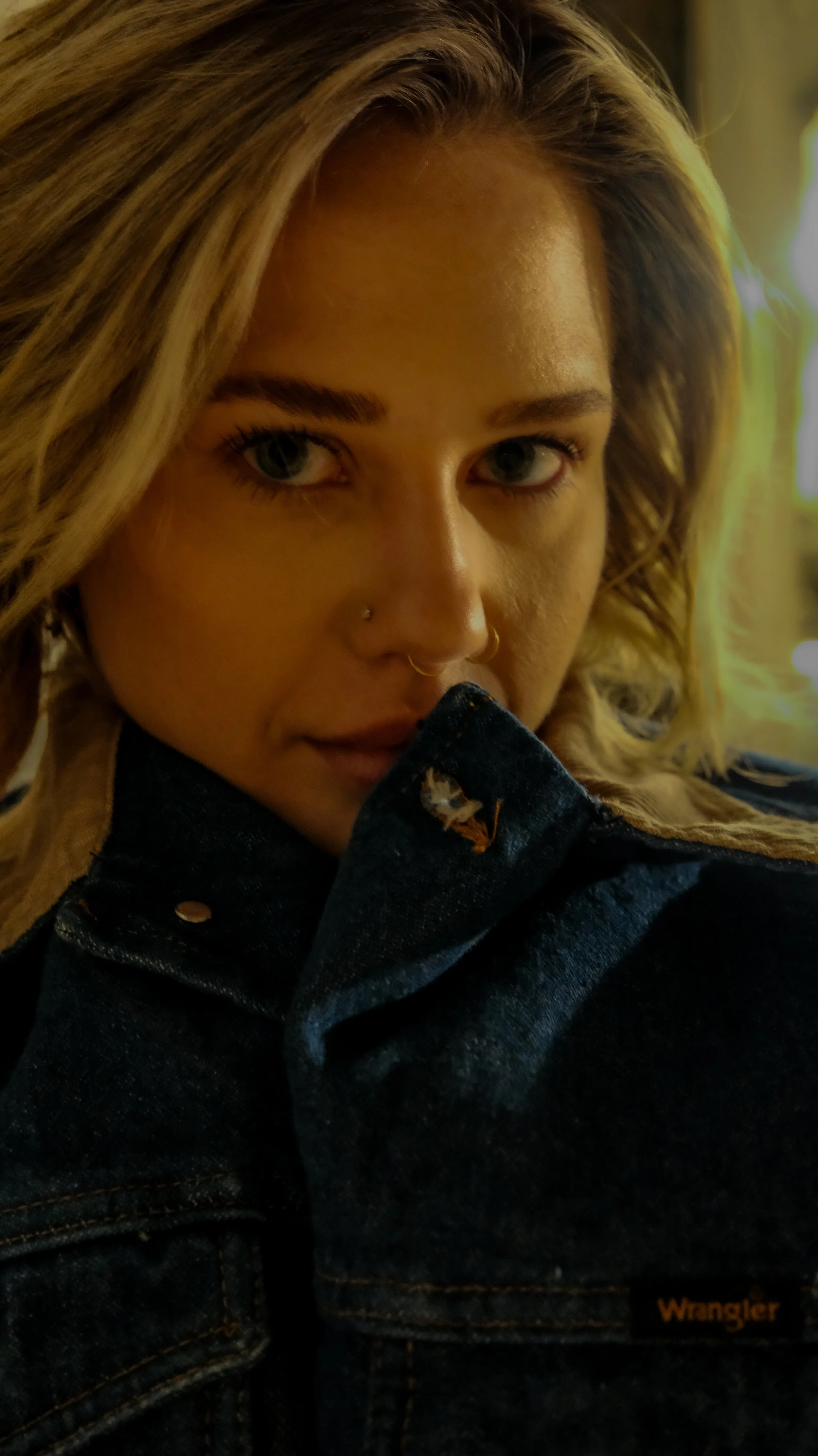 A woman with blonde hair, blue eyes, and piercings near her nose, wearing a dark denim jacket with an orange 'Wrangler' label, looking into the camera with a serious expression.