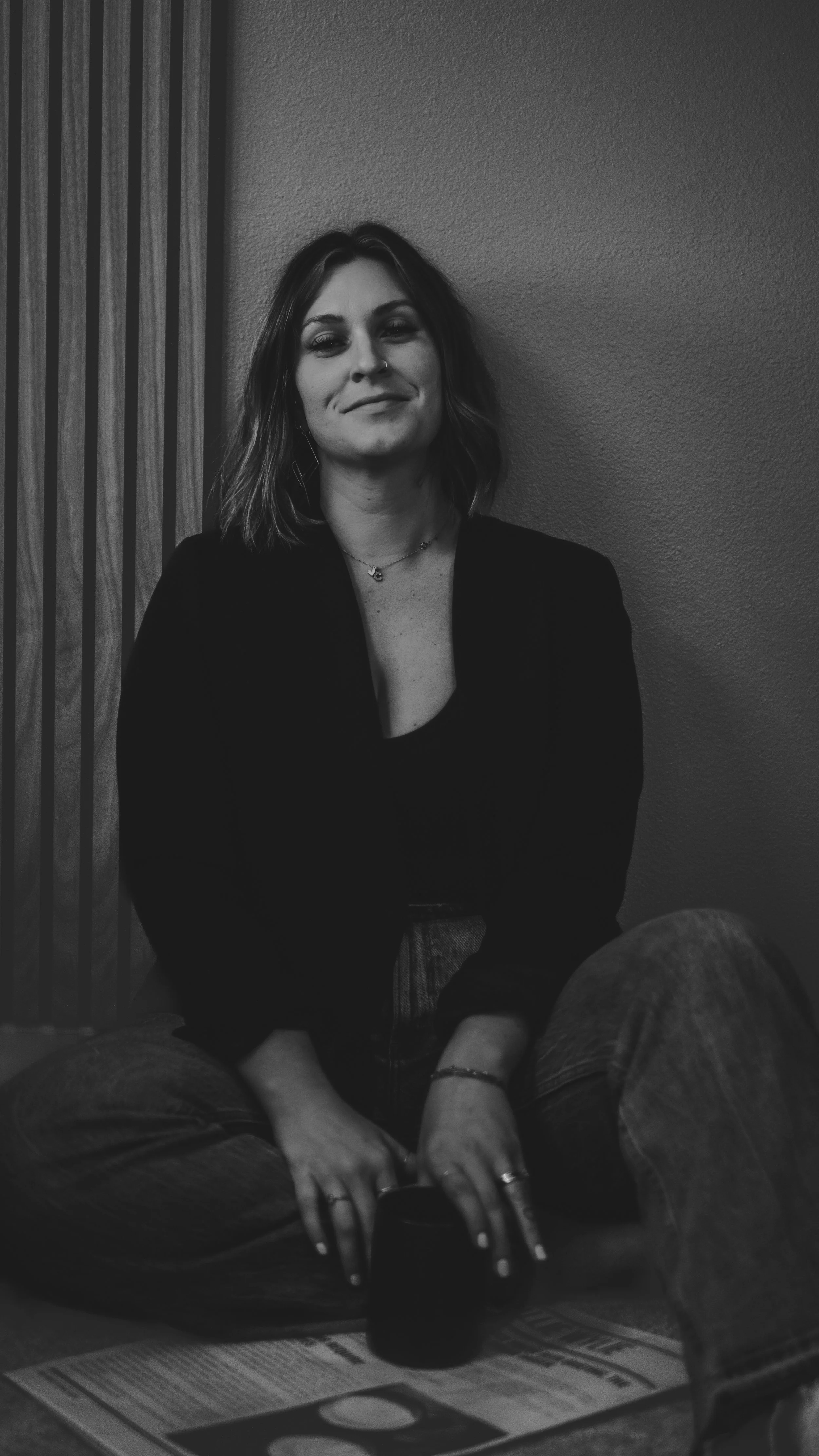 A woman with shoulder-length hair, sitting cross-legged on the floor, holding a mug, with a relaxed expression, wearing a dark jacket and jeans, against a plain wall.