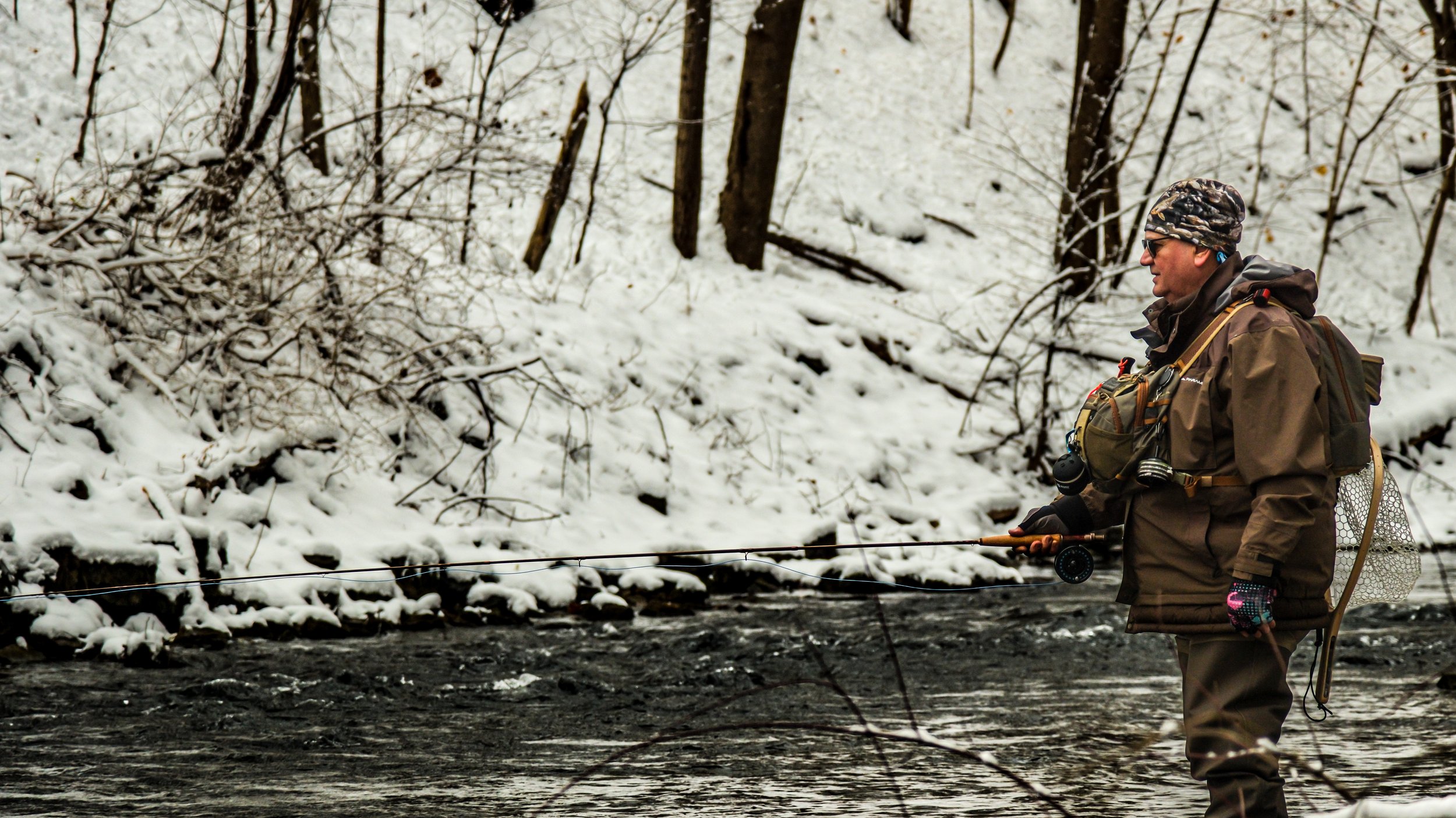Man fishing in a river during winter, surrounded by snow-covered trees.