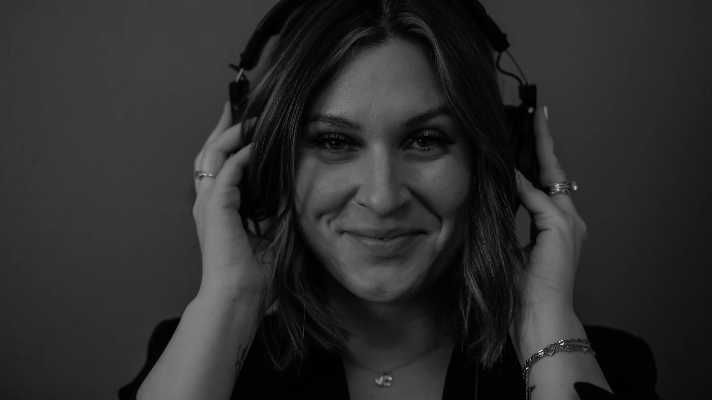 A woman with shoulder-length hair, smiling, wearing headphones, and wearing rings, bracelets, and a necklace.