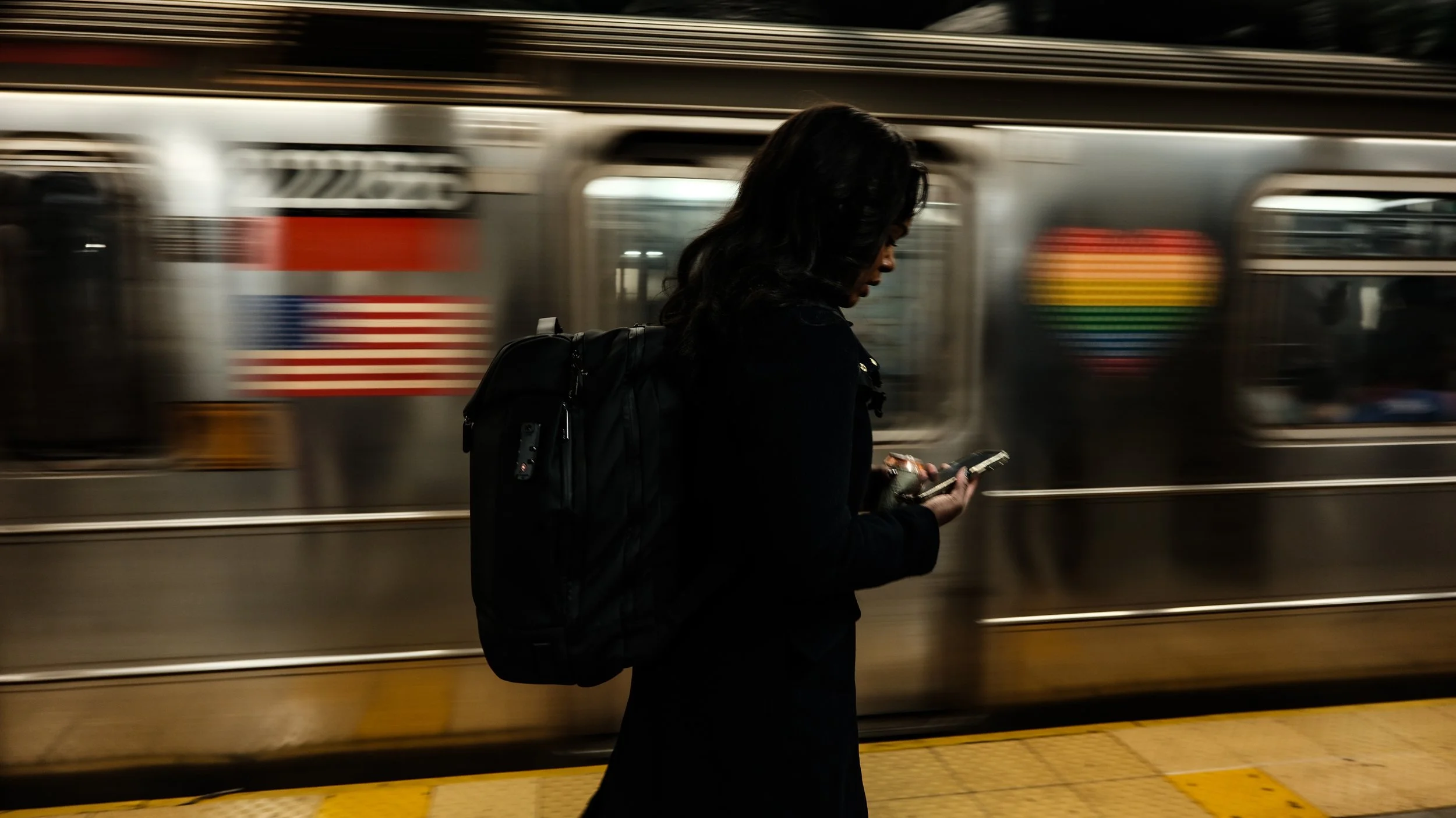 Woman with black backpack waiting on subway platform, looking at her phone, with a moving subway train in the background.