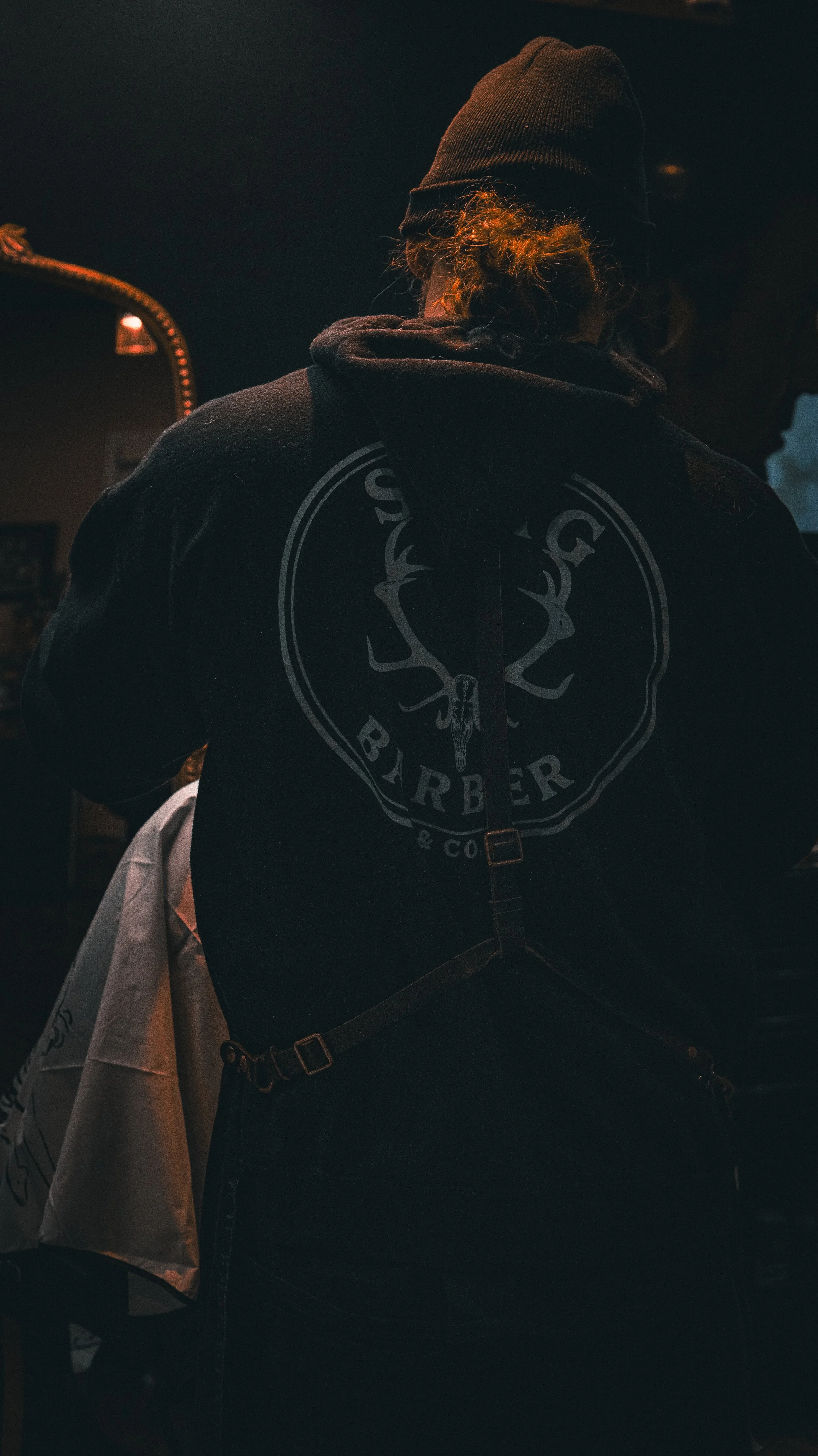 Person with curly hair wearing a black beanie and black hoodie with a barber logo on the back, standing in a dimly lit barbershop.