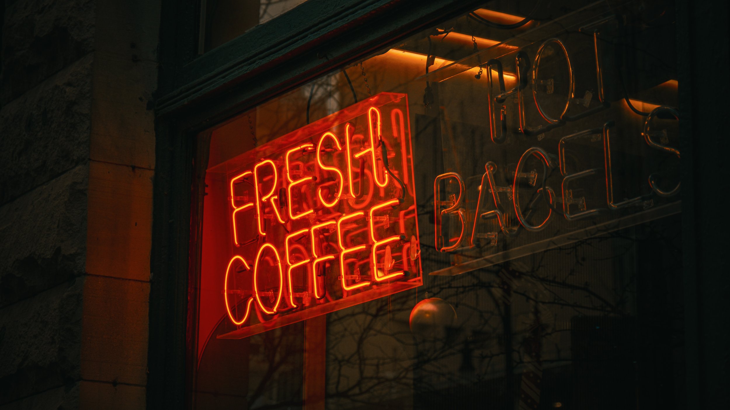 Neon sign in a shop window displaying 'FRESCH CAFÉ' and 'RACHEL' in orange-red glow, with reflections of nearby trees and buildings visible in the glass.