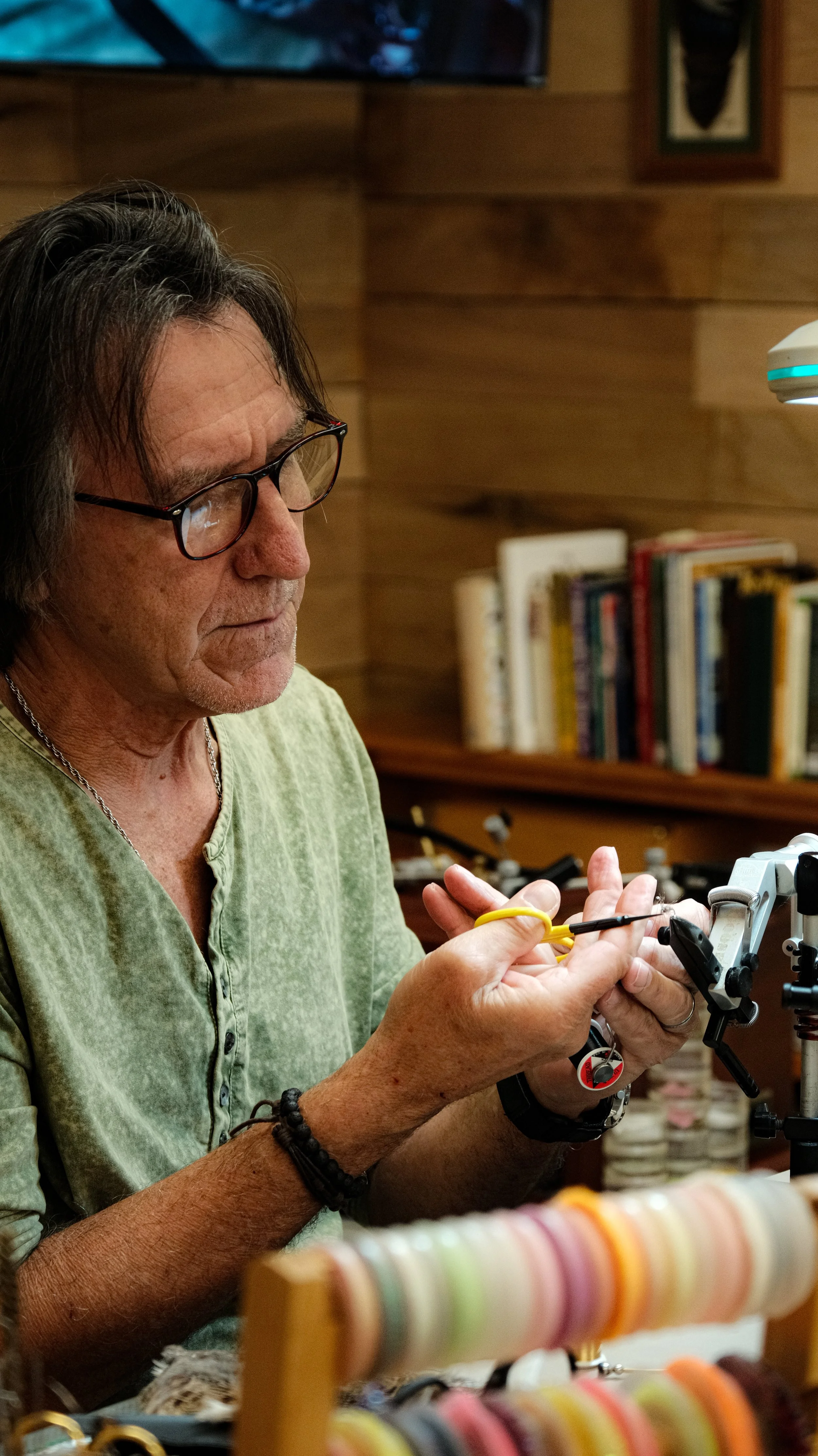 An older man with glasses and long dark hair working on a jewelry or craft project, with colorful beads or thread in the foreground and a bookshelf in the background.