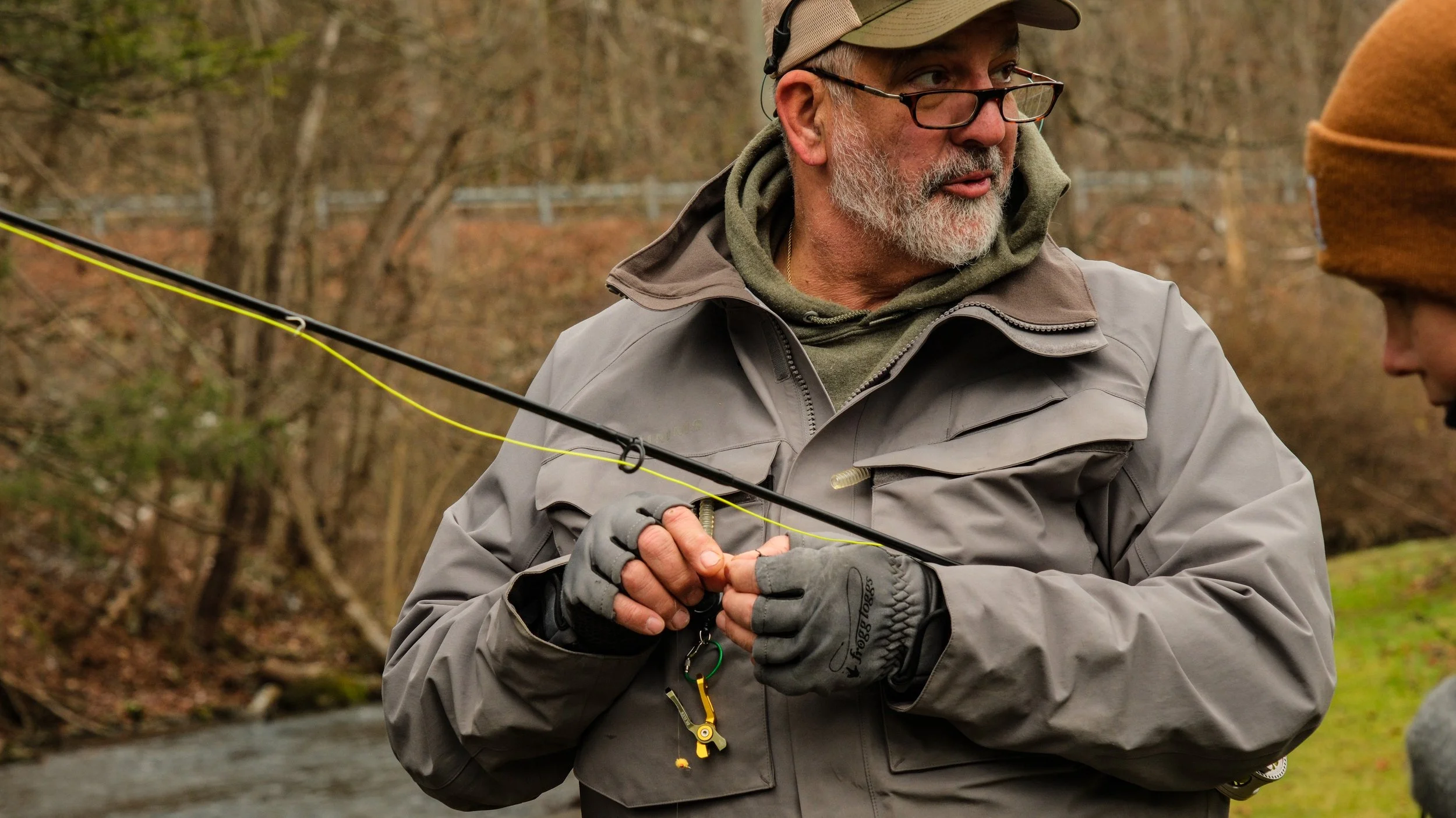 Older man with gray beard wearing glasses, a cap, and outdoor clothing, holding a fishing rod and talking to a woman in a brown beanie, next to a stream in a wooded area.
