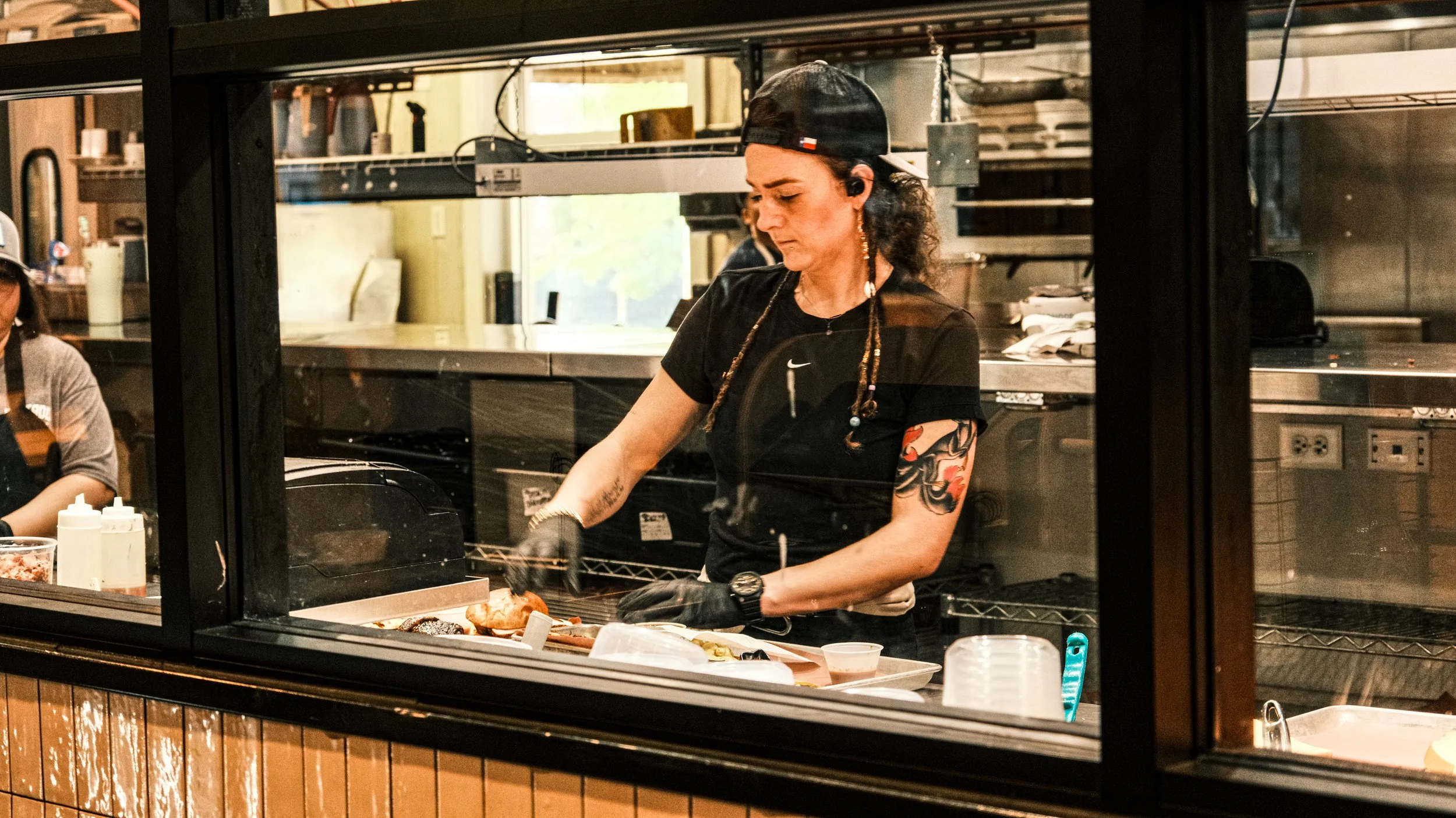 A woman wearing a black cap, black shirt, and black gloves preparing food behind a glass window in a kitchen or restaurant environment.