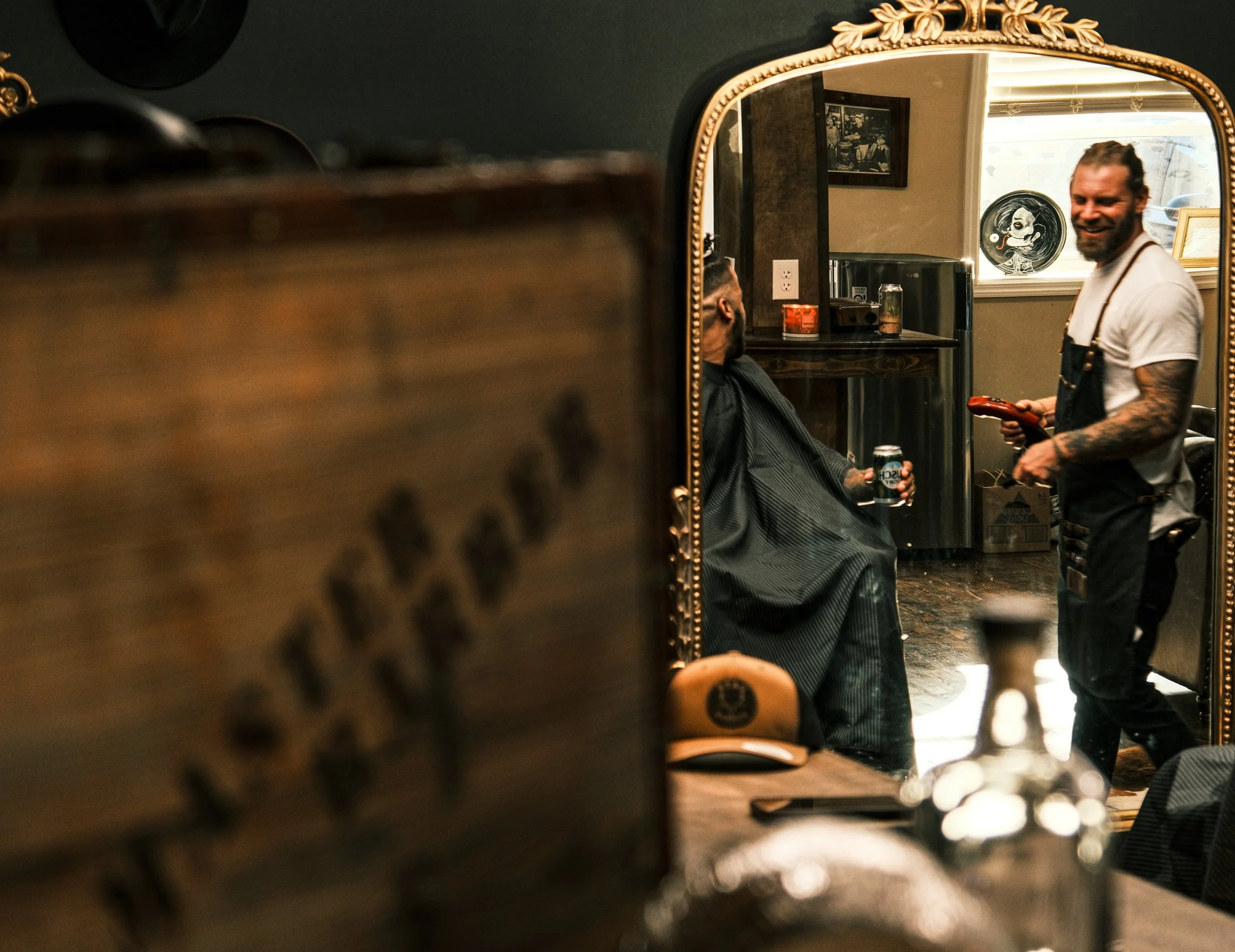 A barber shop with a large ornate mirror showing a man with tattoos in a barber chair holding a drink, and a smiling barber in black apron holding a spray bottle, with tools and decor visible in the background.