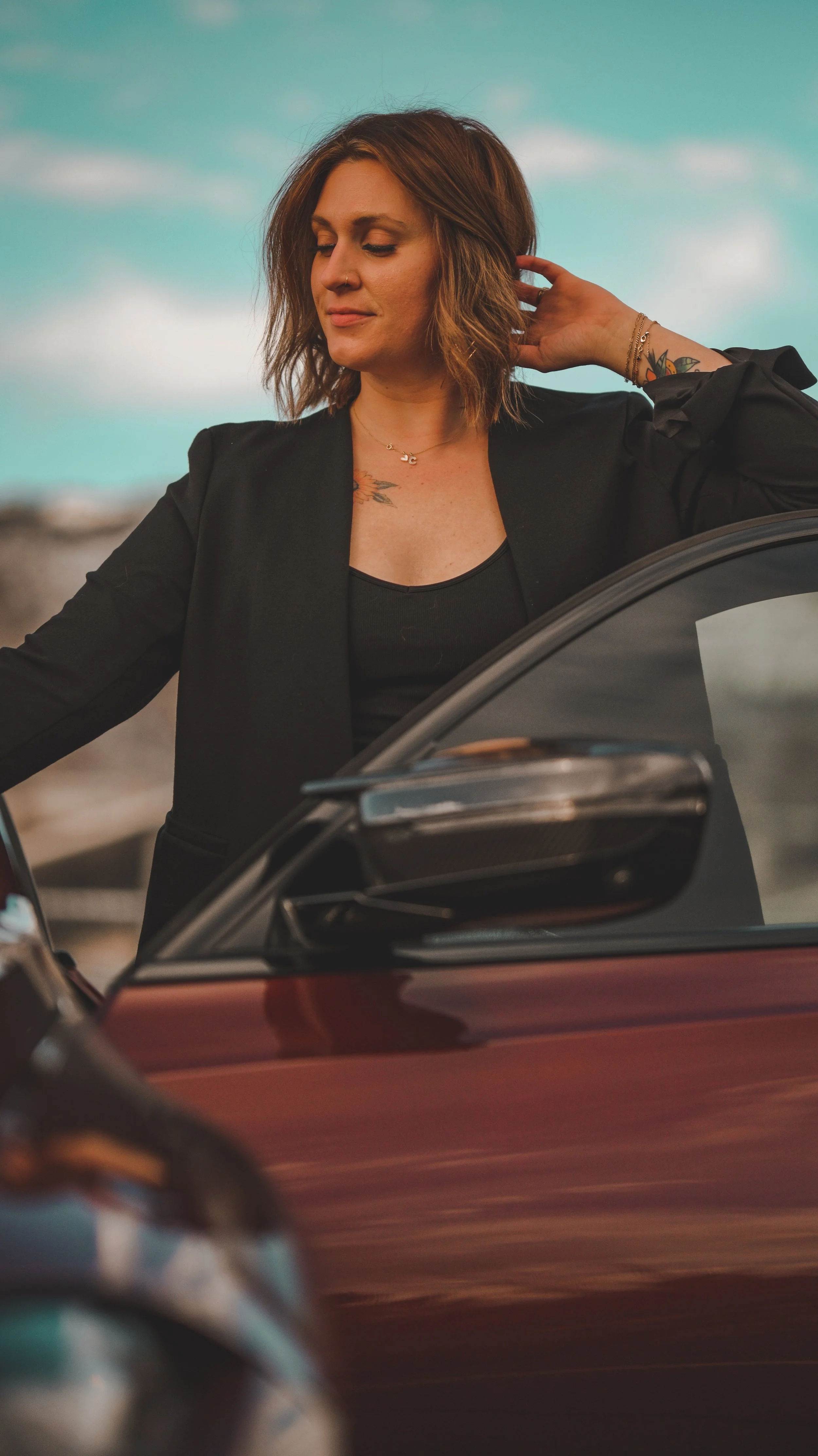 A woman with brown, shoulder-length hair standing next to a red car, touching her neck with her eyes closed and a slight smile, outdoors with a cloudy sky in the background.