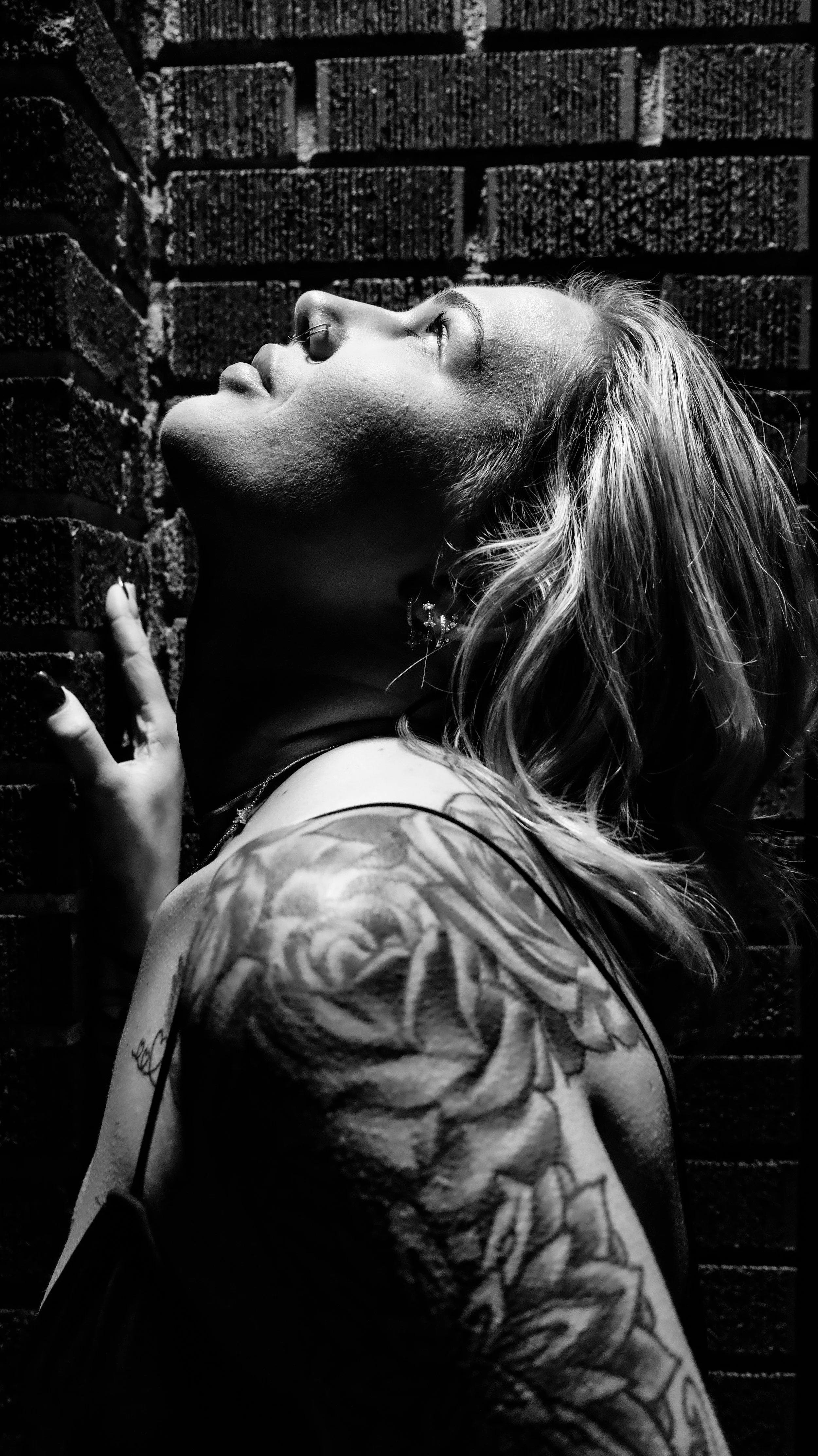 A black and white photo of a woman with tattooed arm and earrings, looking upward against a brick wall.