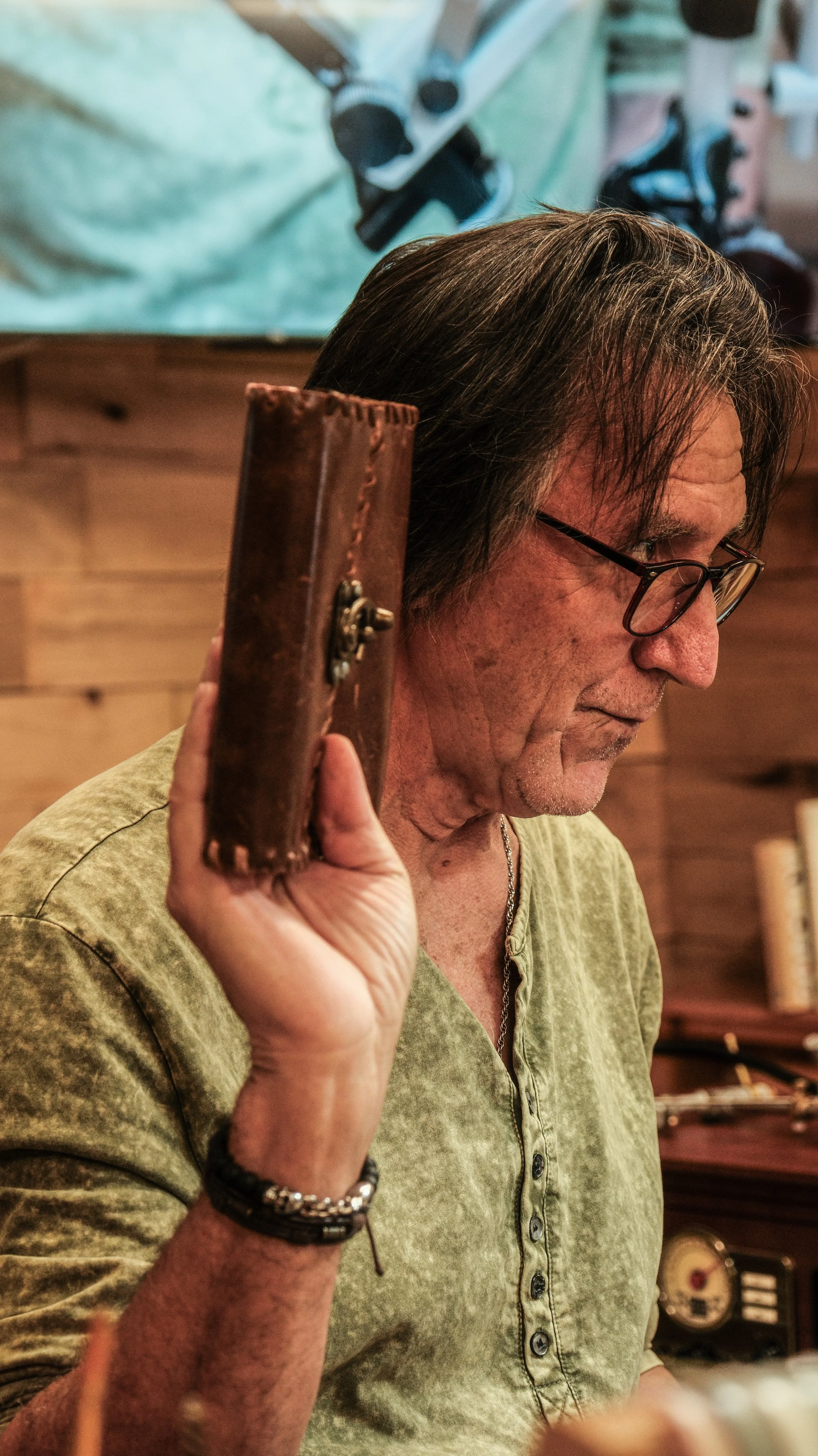 A man with brown hair, glasses, and a green shirt holding a leather wallet with a latch to his ear in a wood-paneled room.