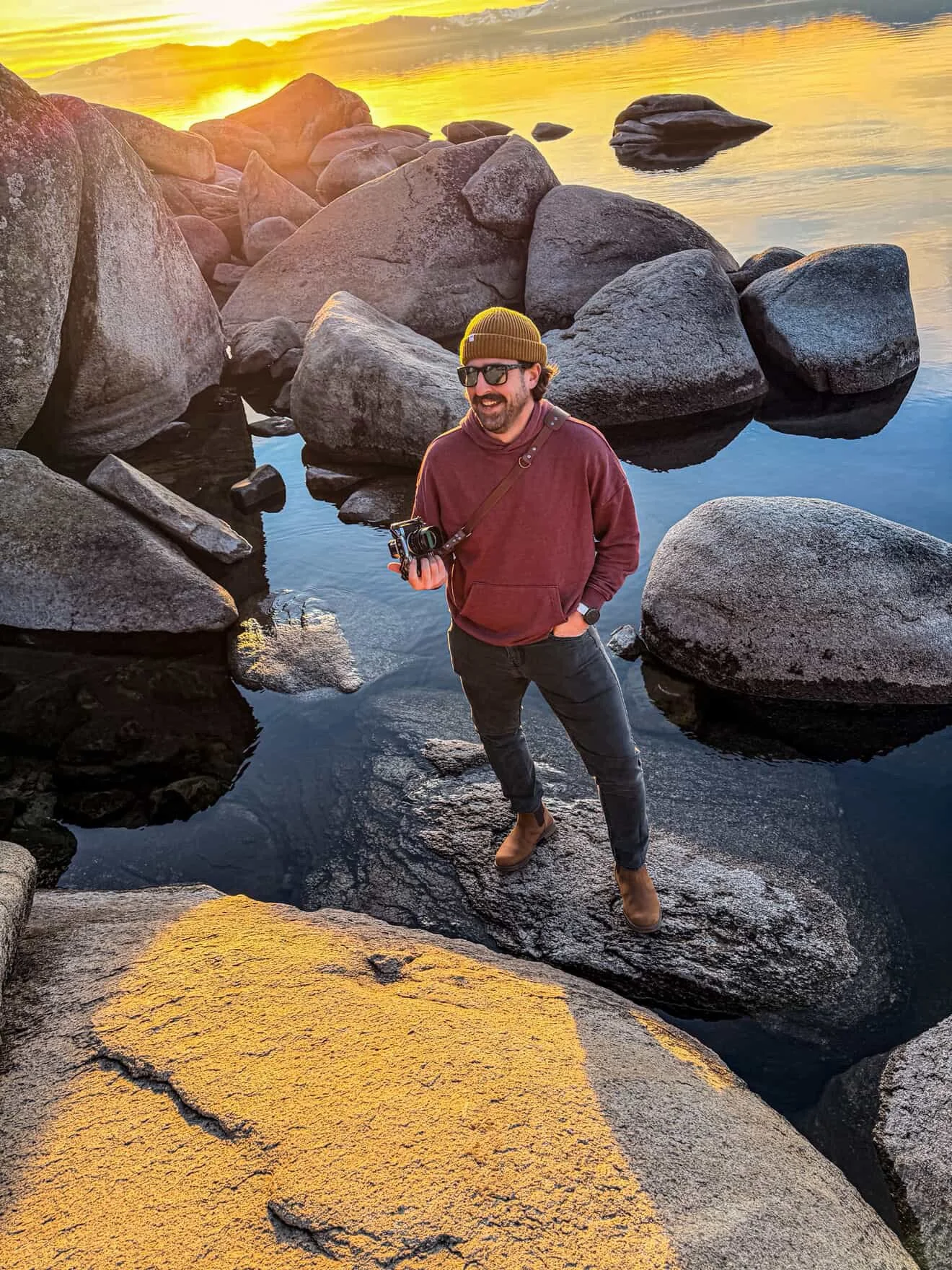 A man with sunglasses, a brown beanie, a maroon hoodie, and black pants stands on a large rock in a body of water surrounded by large boulders during sunset.