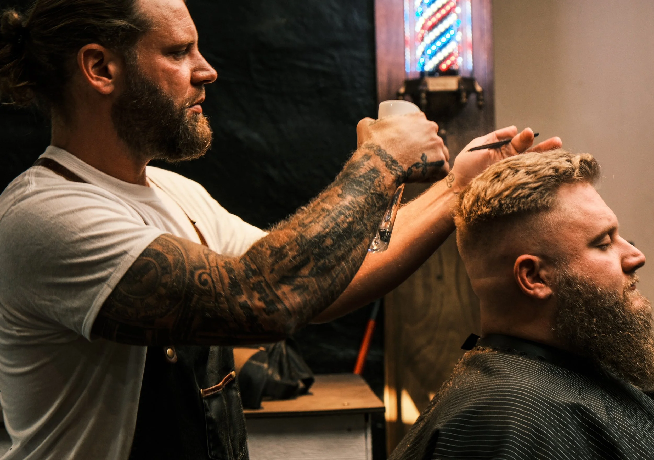 Barber cutting a man's hair in a salon, using scissors and a comb, with a tattooed barber on the left side.