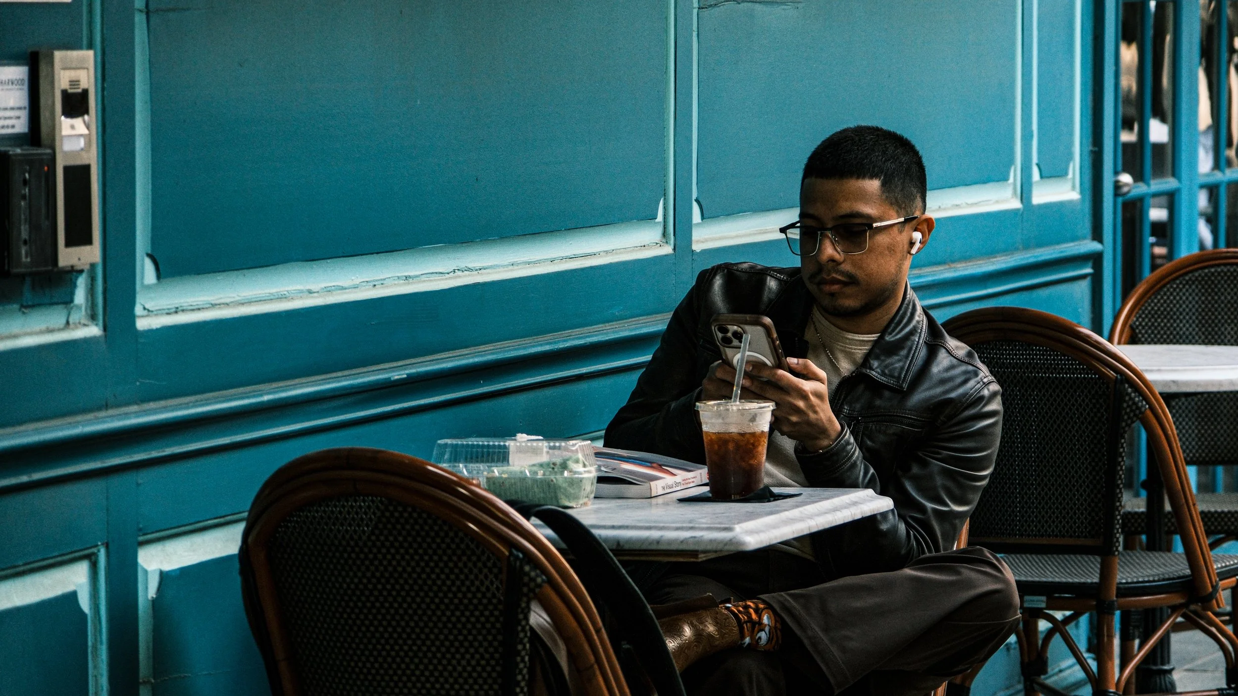 A man wearing glasses, a leather jacket, and wireless earbuds sitting alone at a table in a cafe, looking at his phone, with a cold drink and food containers on the table.
