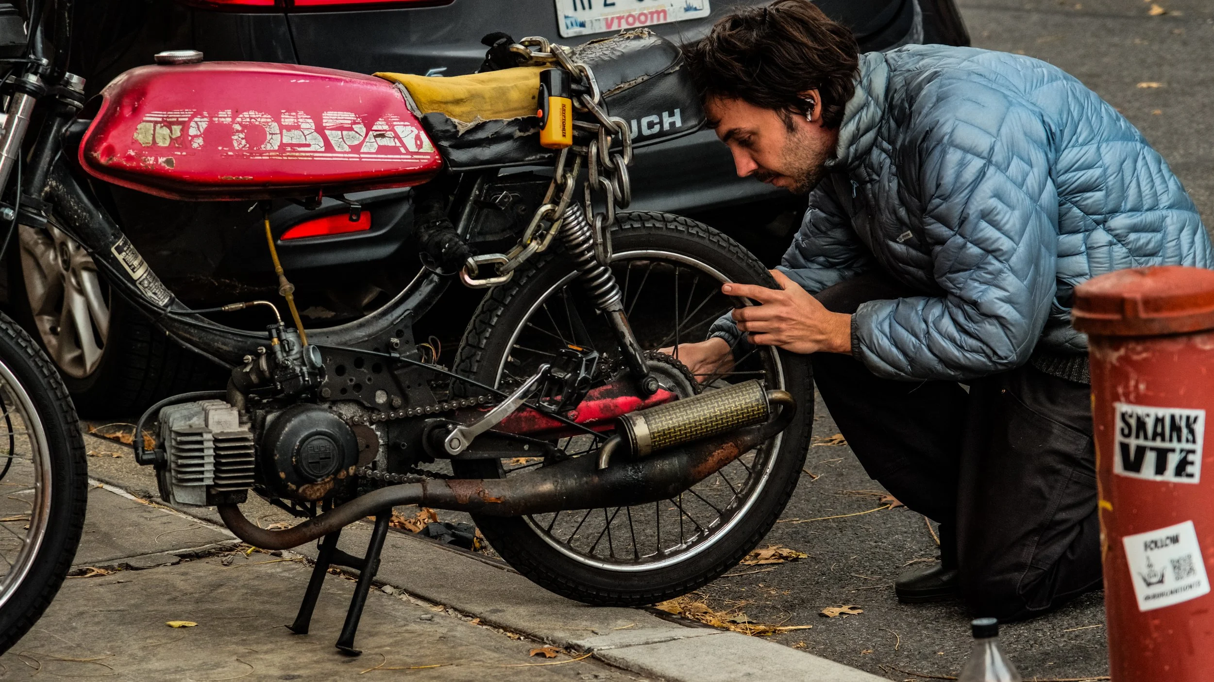 A man in a blue quilted jacket working on a vintage motorcycle parked on a city street.