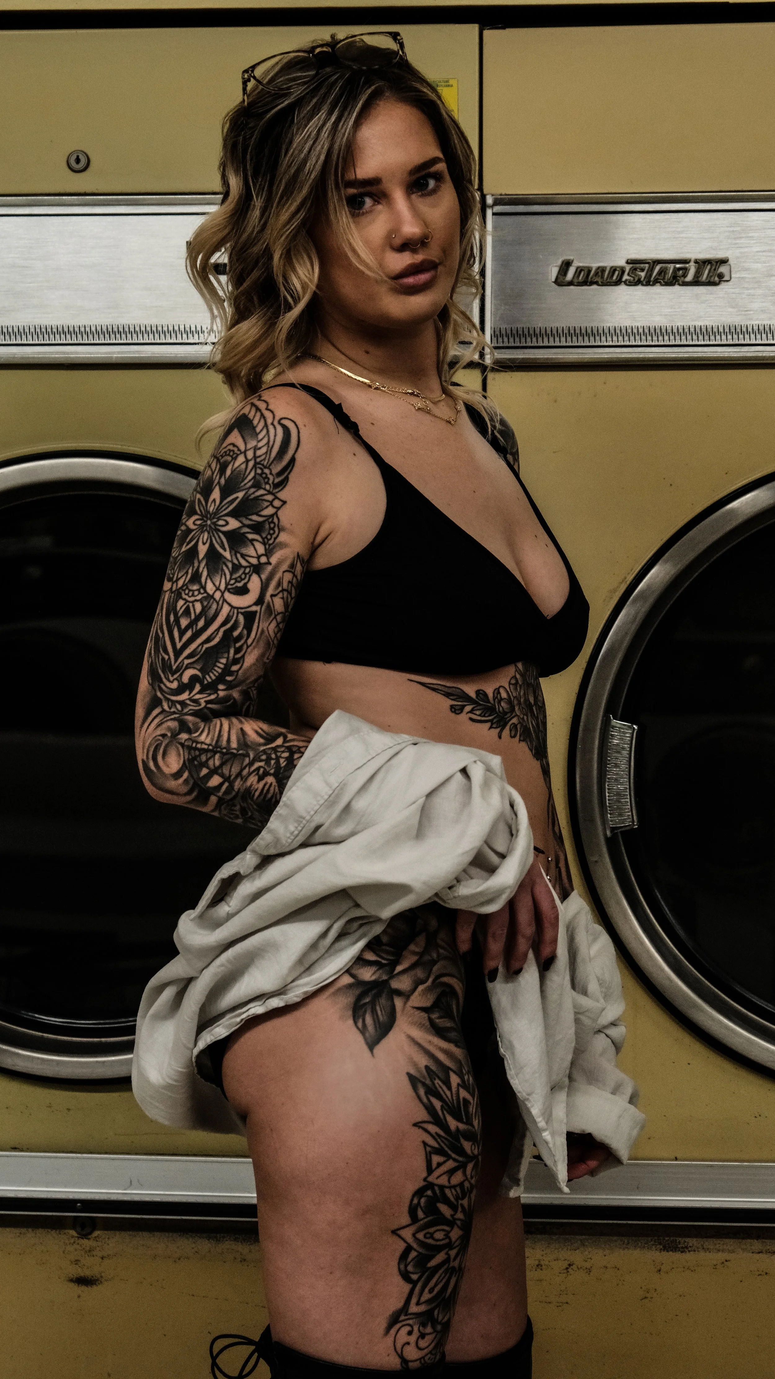Young woman with tattoos holding a white shirt in front of a laundromat dryer.