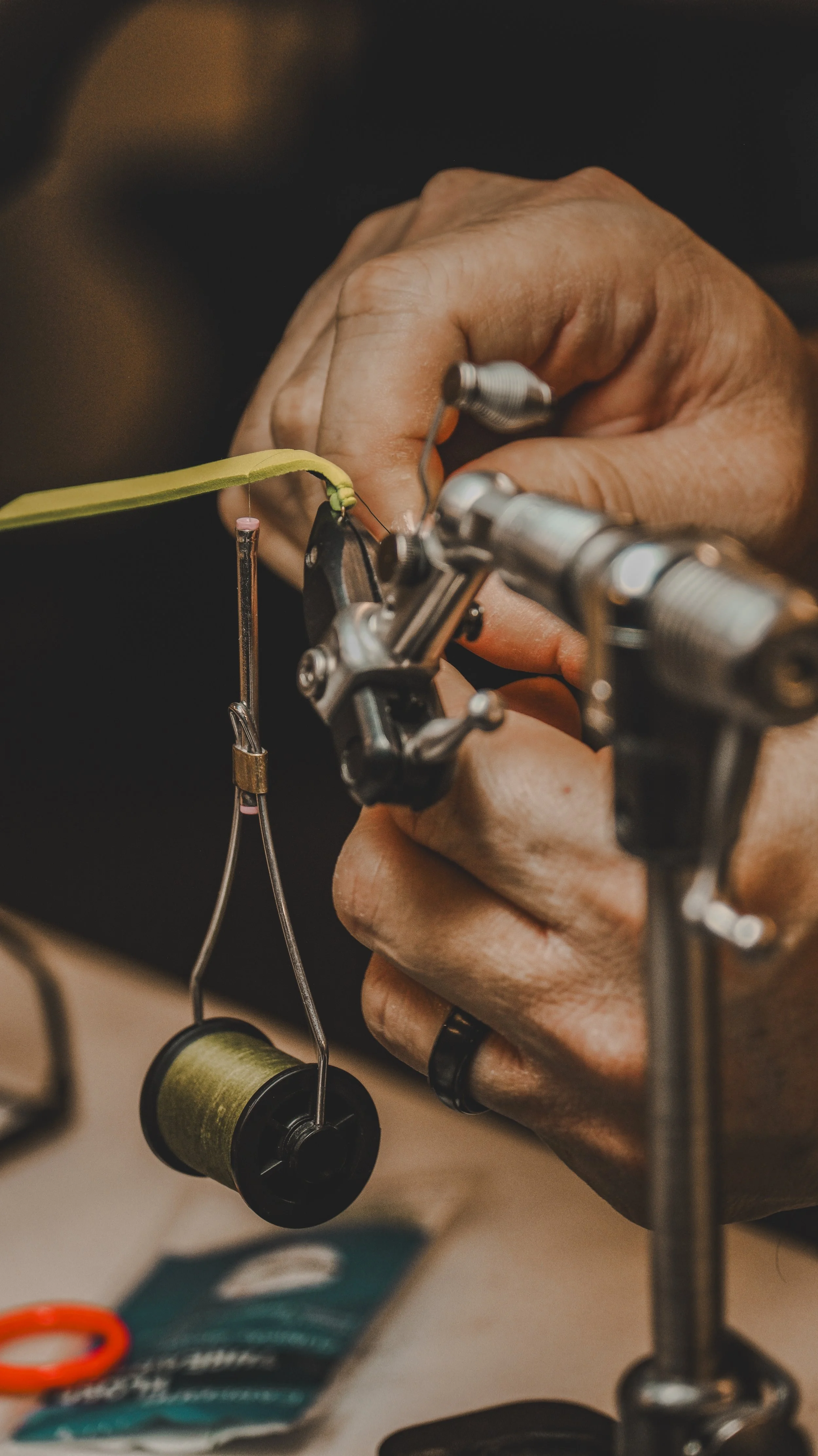 Close-up of hands using a fly tying tool to attach a pink and green hook to a fishing lure, with a spool of green thread hanging nearby.