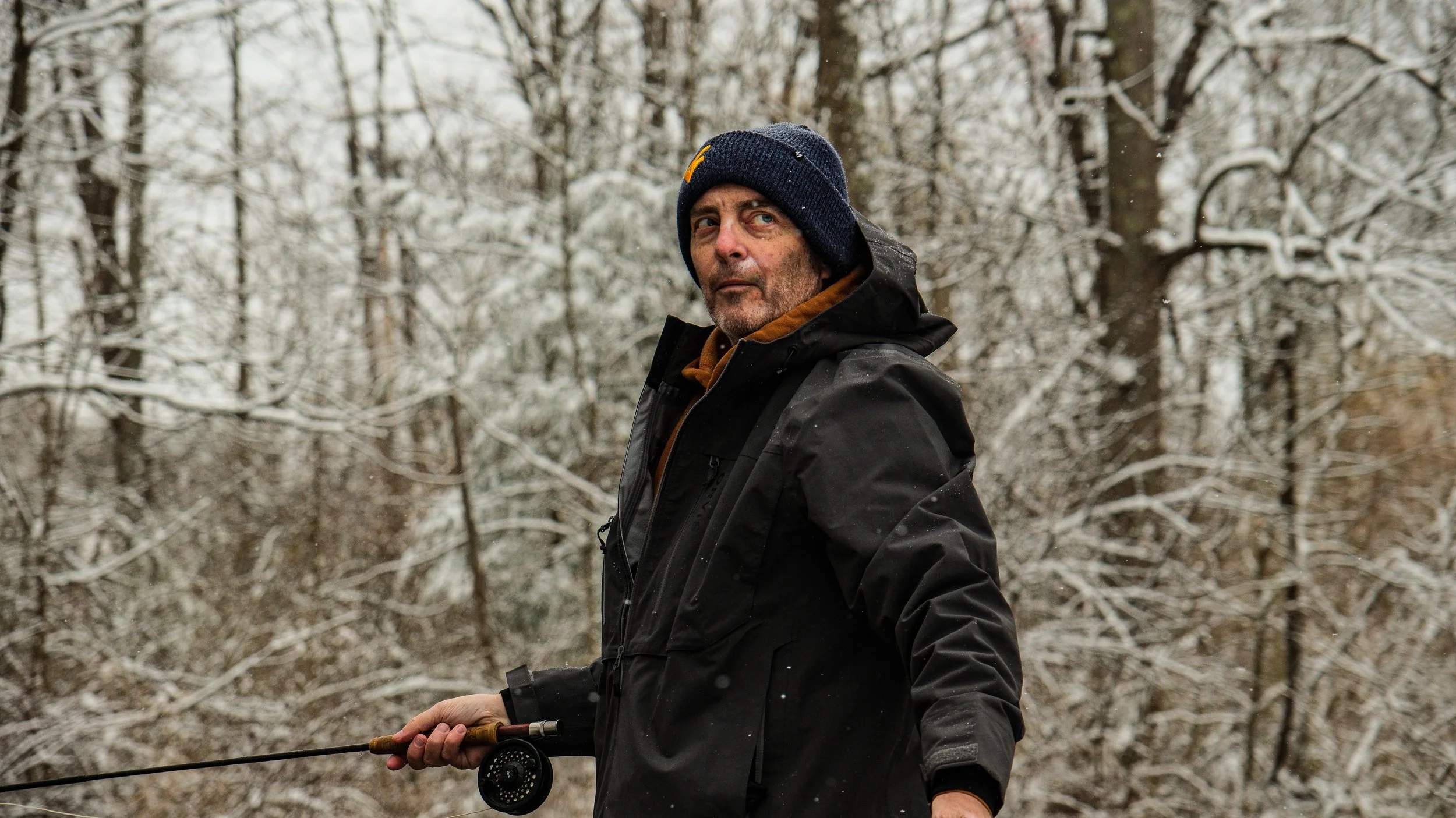 A man in a black jacket and navy blue beanie holds a fishing rod in a snow-covered forest.