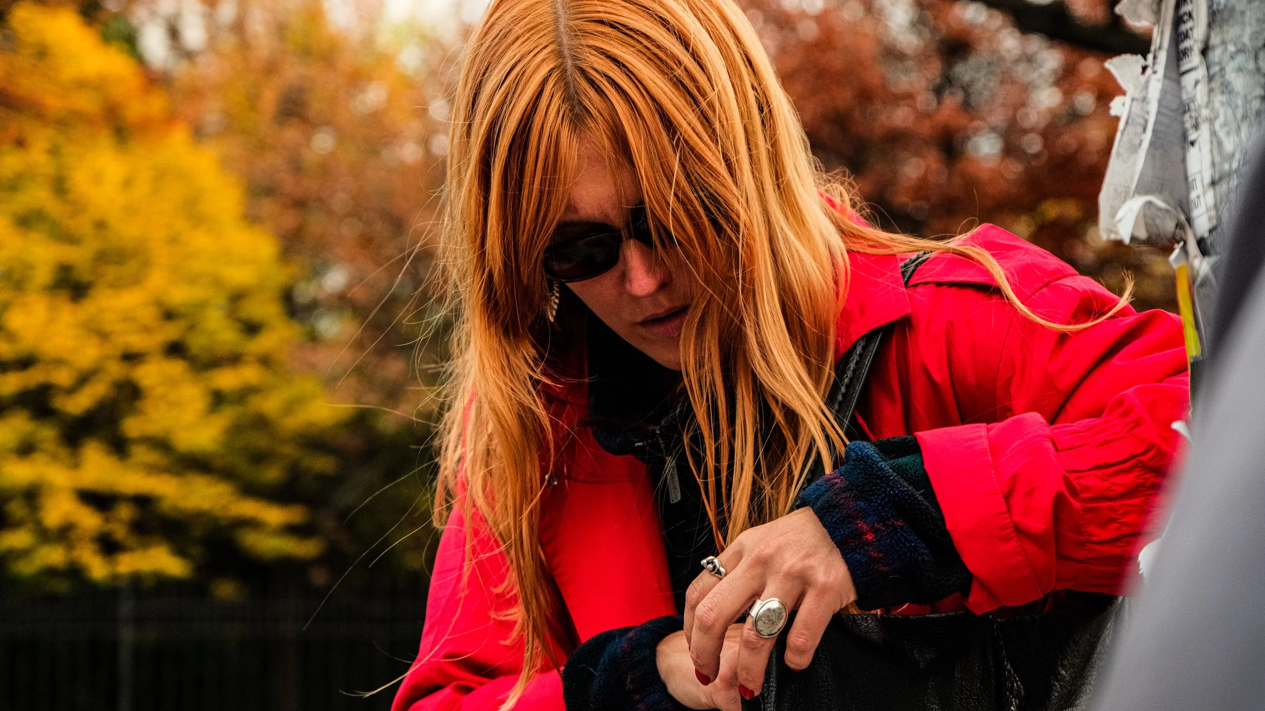 A woman with long red hair wearing dark sunglasses and a red jacket is outdoors, looking down and working on something with her hands, with autumn trees in the background.