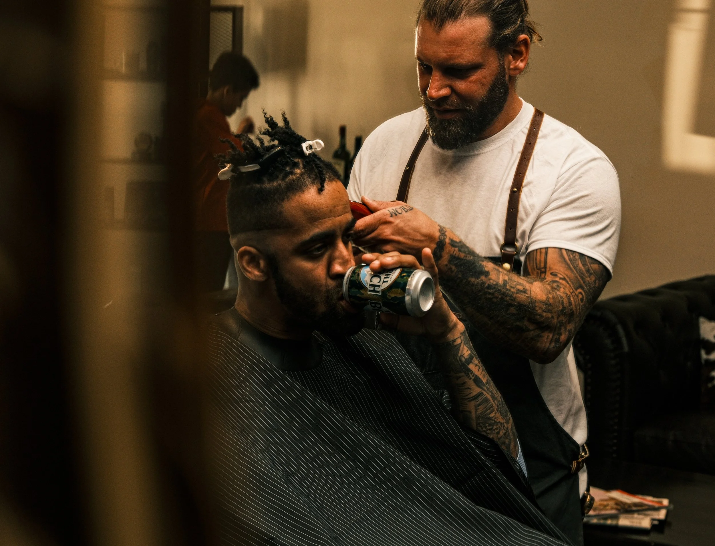 A man getting a haircut at a barbershop, sipping from a can, while a barber cuts his hair. The barber has tattoos on his arms and is focused on his work.