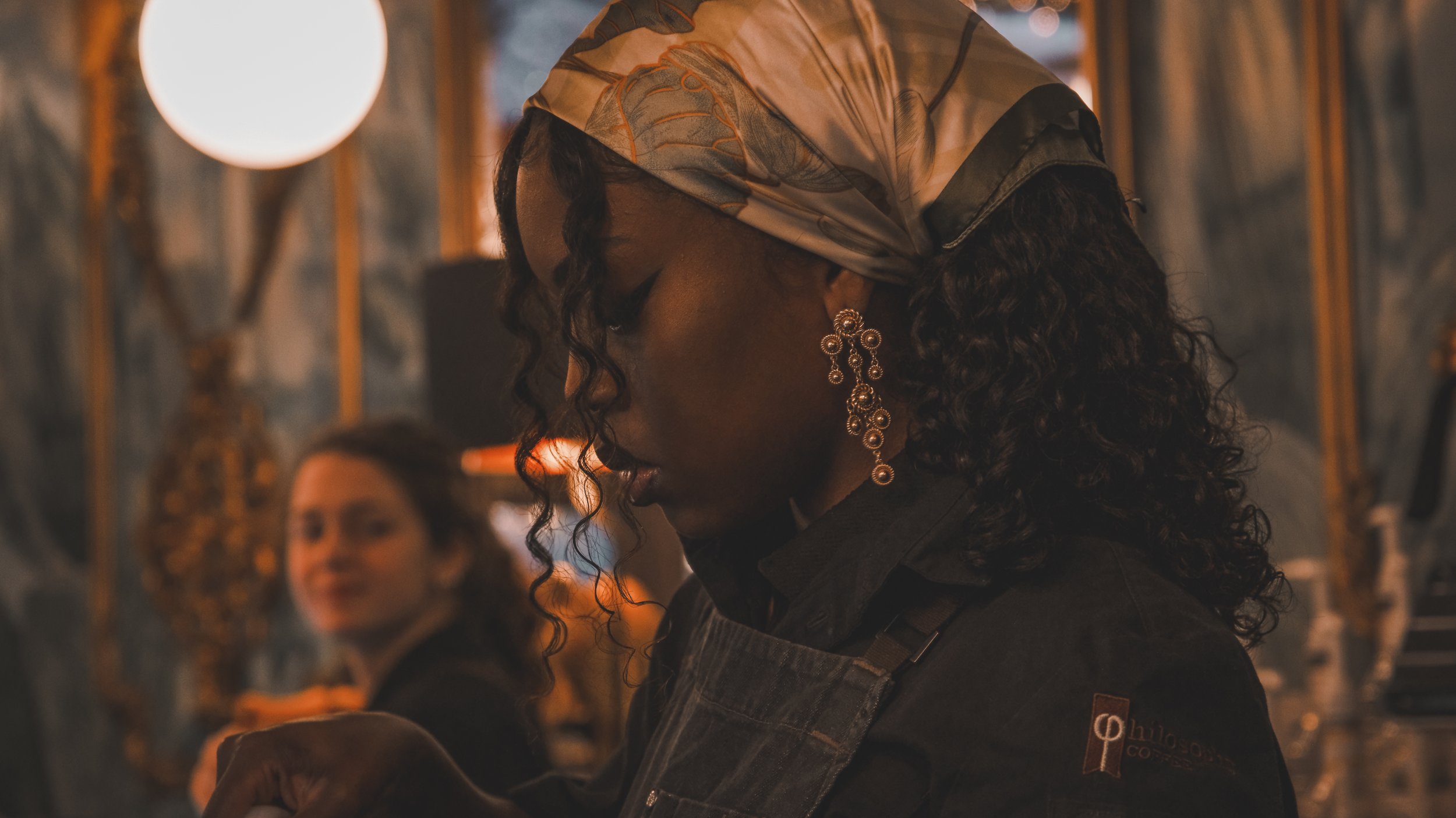 A woman with curly black hair wearing a colorful headscarf, statement earrings, and a dark apron inside a restaurant or cafe, with another woman blurred in the background.
