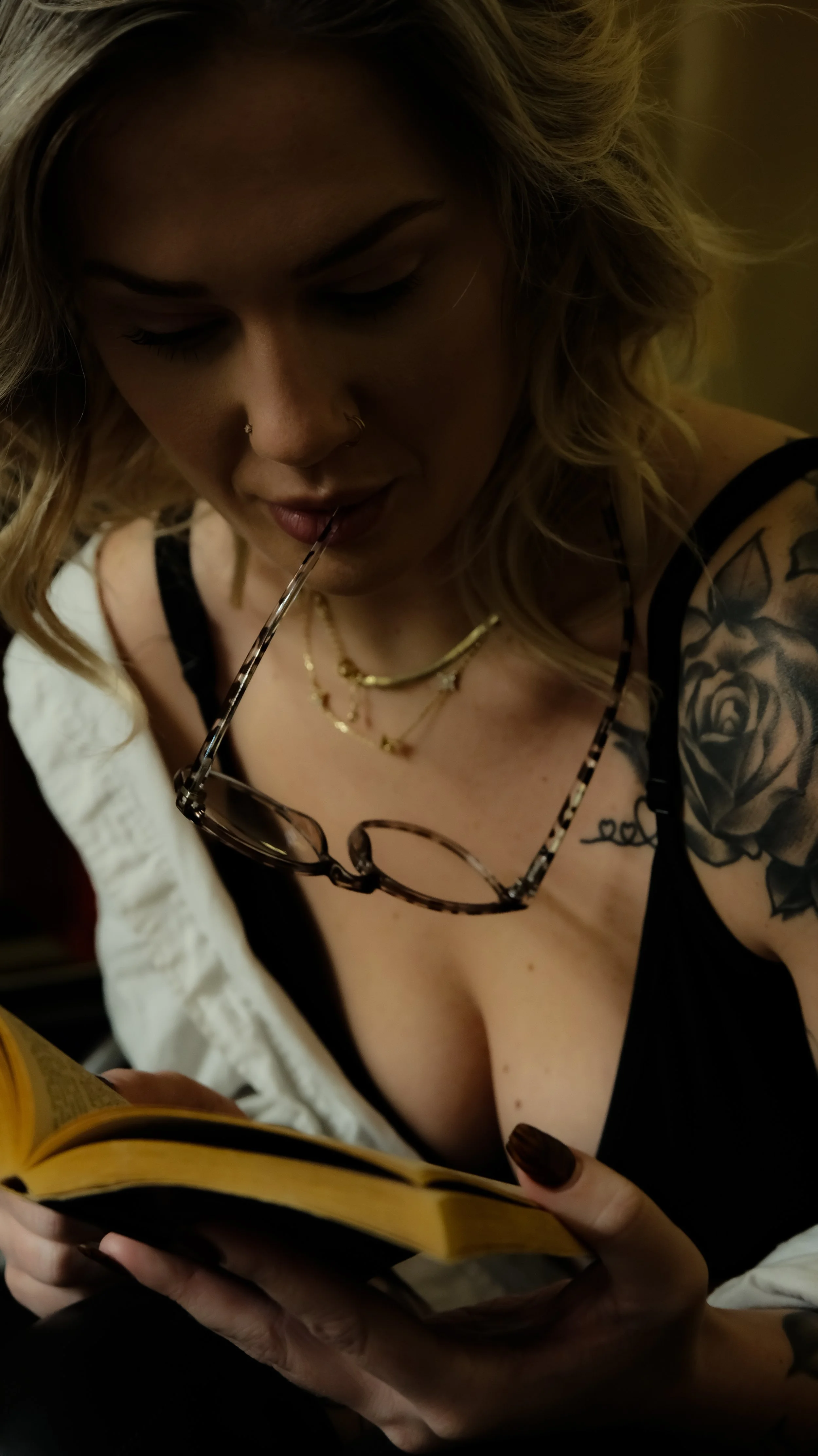 A woman with blonde hair, tattoos on her shoulder, and glasses hanging from her mouth, reading a book or magazine.