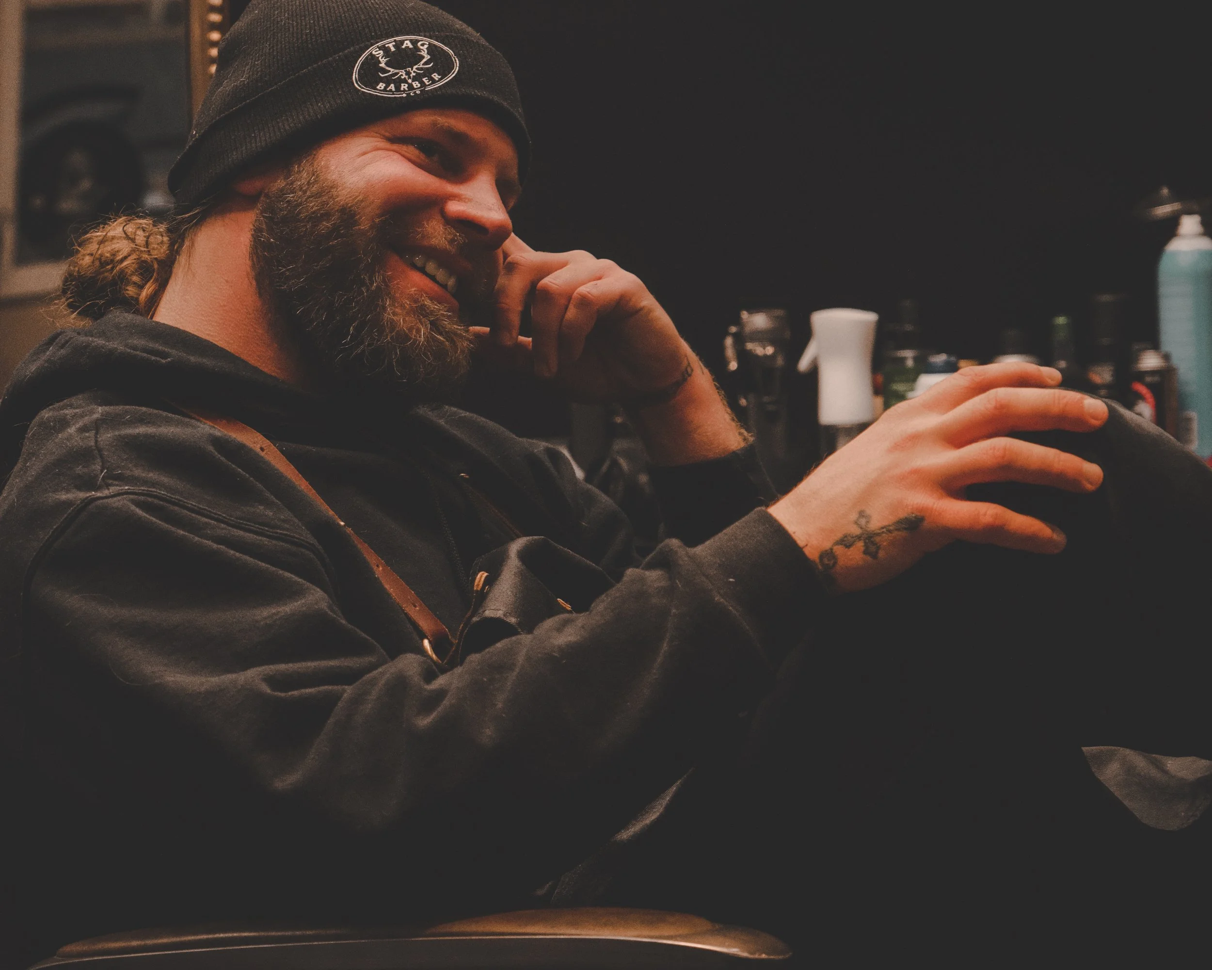 A man with a beard, wearing a black beanie and hoodie, smiling while sitting in a dimly lit indoor setting, with tattooed hand resting on his knee and a cluttered background of toiletries and bottles.