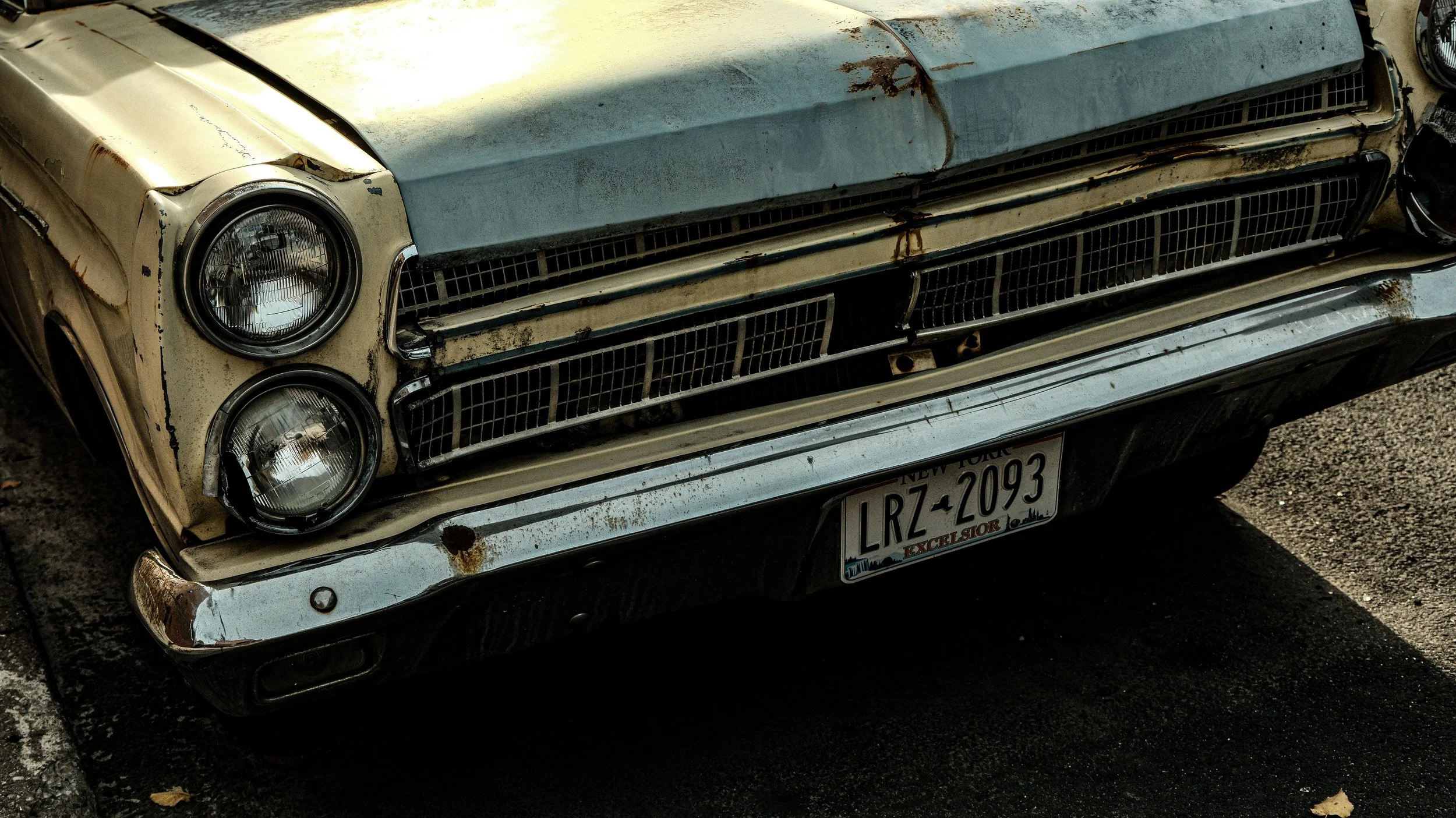 Close-up of an old, rusty vintage car with a worn beige and grey exterior, round headlights, and a New York license plate that reads 'LRZ-2093.' The car appears weathered and parked on a dark paved surface with some leaves.