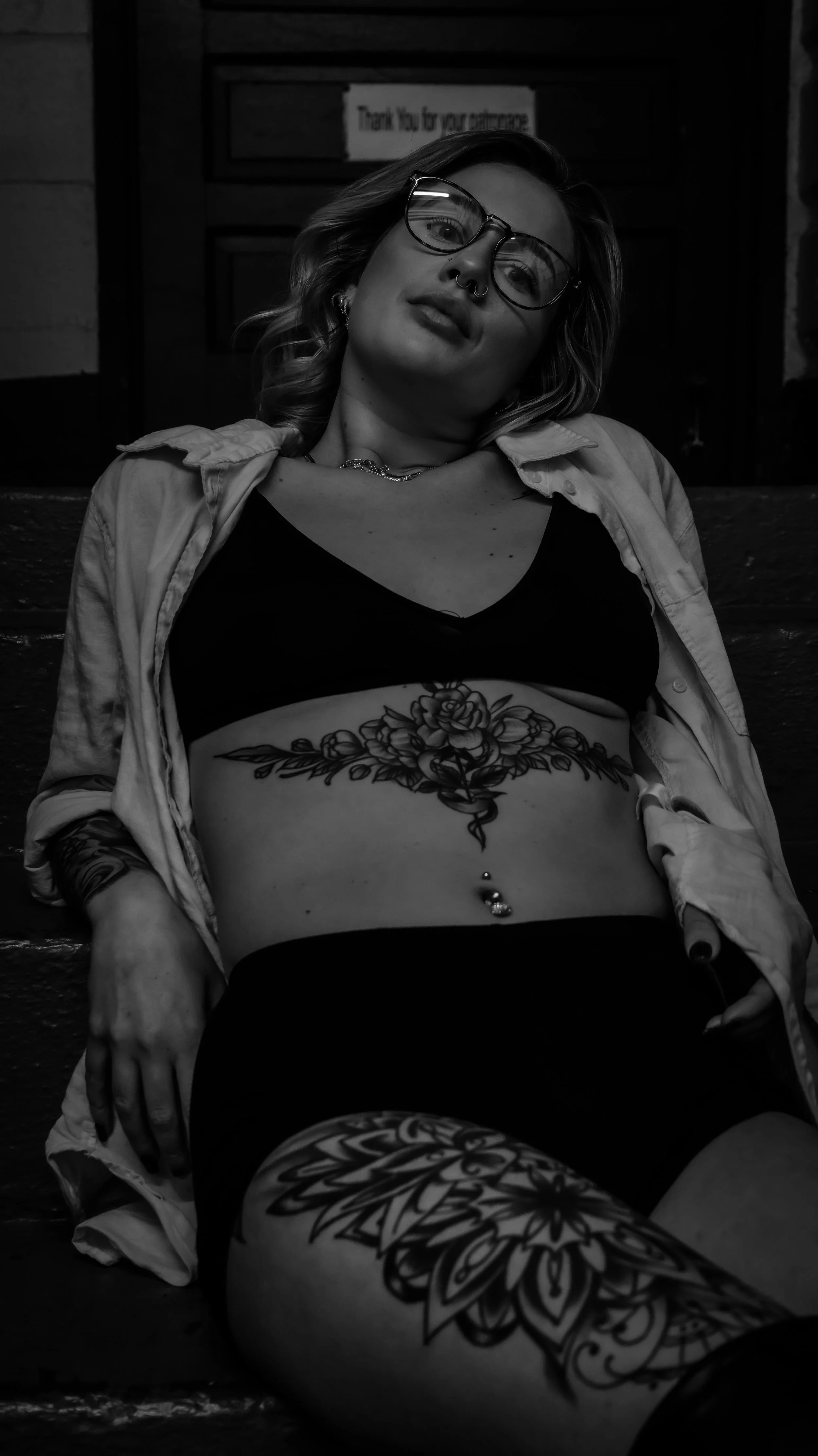 A woman with tattoos on her midsection and thigh, wearing glasses, a bikini top, and an unbuttoned shirt, sitting outdoors at night.